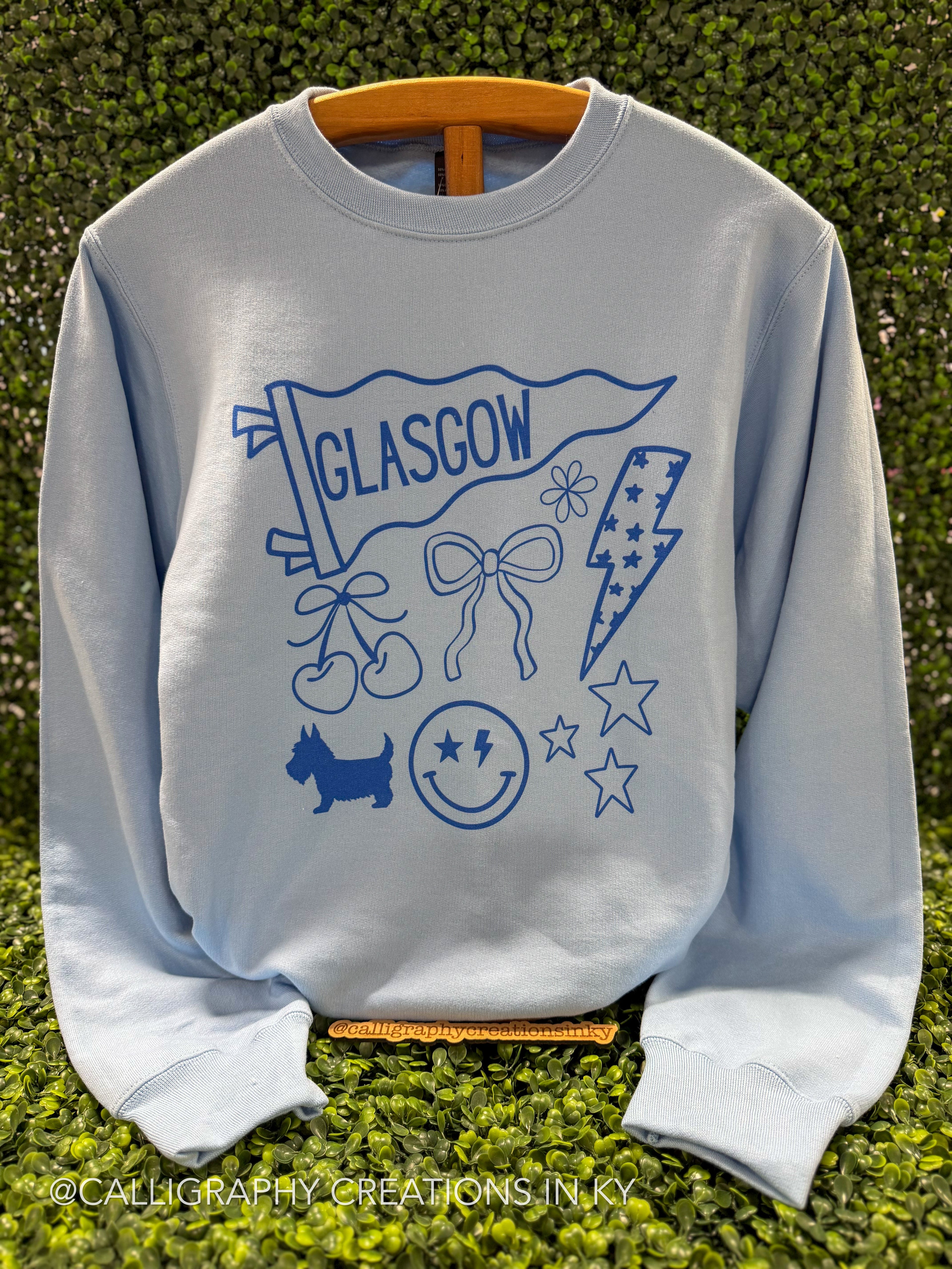 A light blue crewneck sweatshirt featuring a blue graphic print of a Glasgow flag, cherries, bow, lightning bolt, dog, smiley face, and stars, displayed on a wooden hanger against a green hedge background.