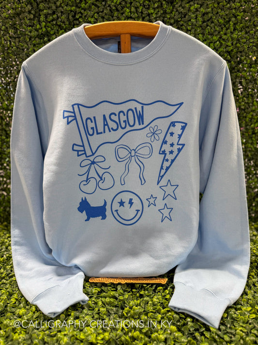 A light blue crewneck sweatshirt featuring a blue graphic print of a Glasgow flag, cherries, bow, lightning bolt, dog, smiley face, and stars, displayed on a wooden hanger against a green hedge background.