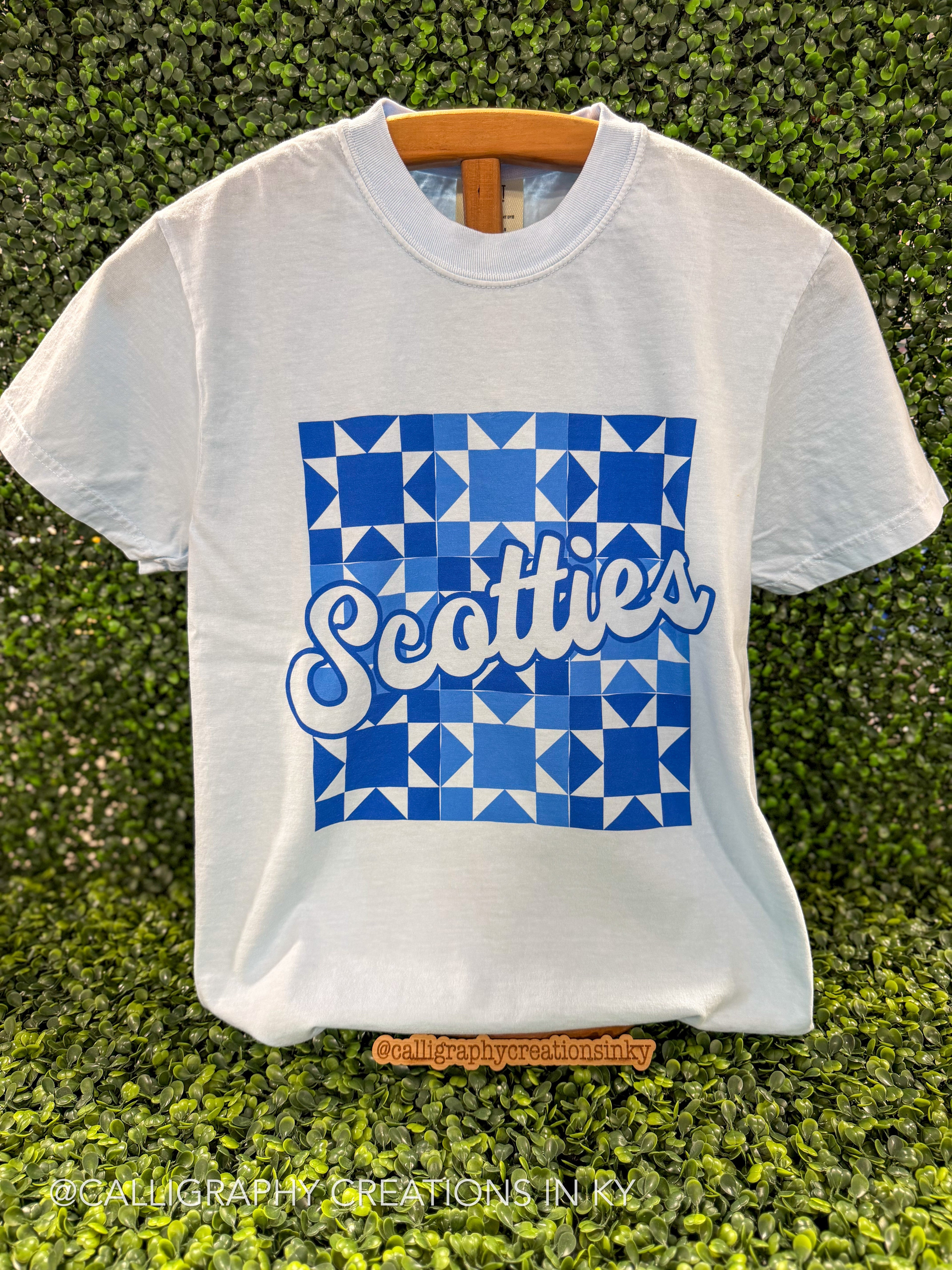A white short-sleeve t-shirt with a blue geometric pattern and the word 'Scotties' in white script, displayed on a wooden hanger against a green hedge background.