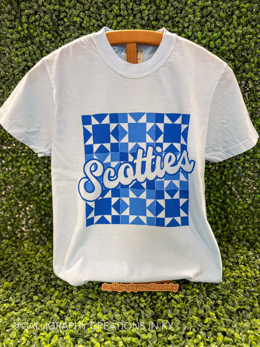 A white short-sleeve t-shirt with a blue geometric pattern and the word 'Scotties' in white script, displayed on a wooden hanger against a green hedge background.