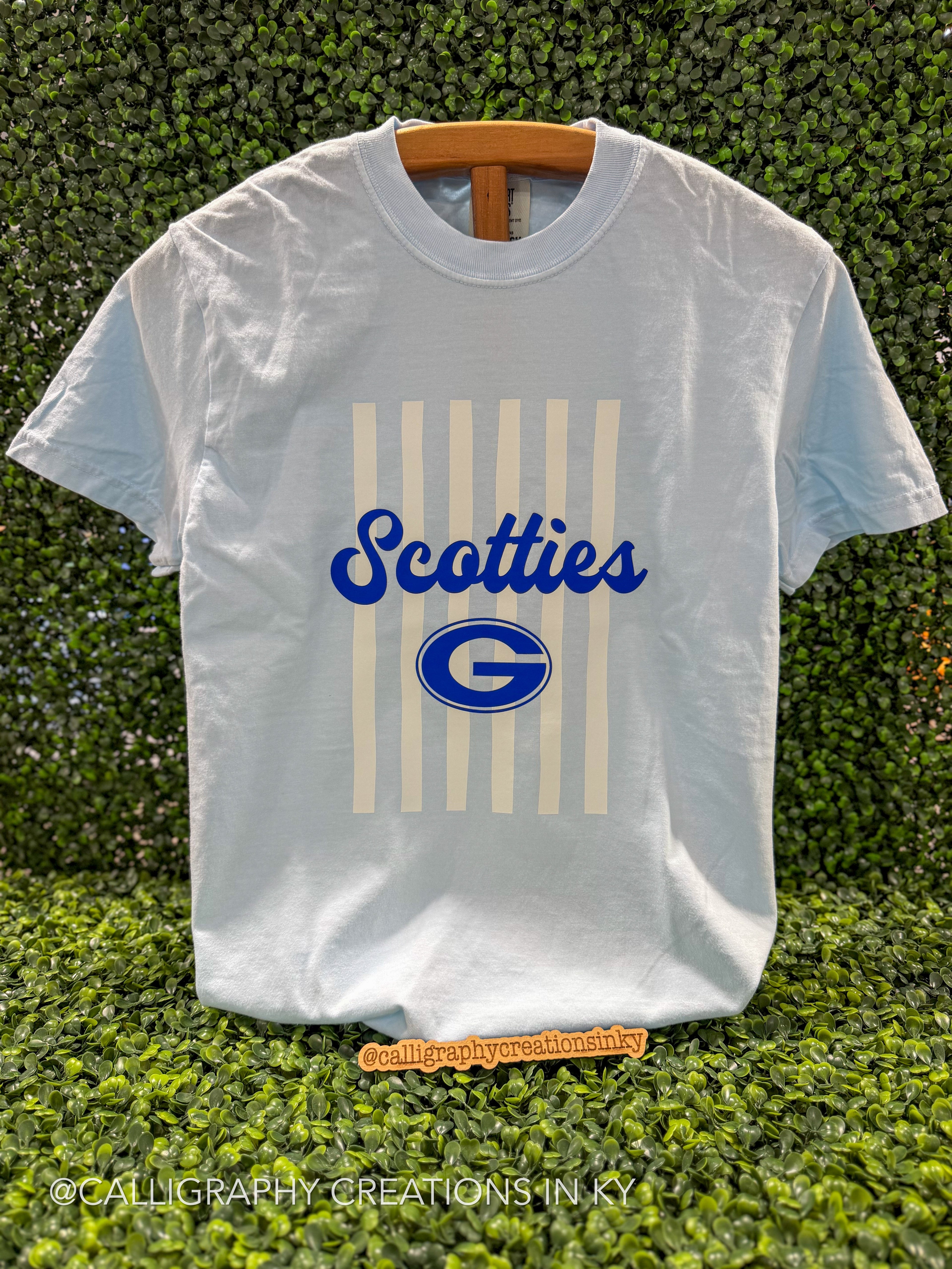 A white short-sleeve t-shirt with a blue and cream graphic print reading 'Scotties' and a large 'G' logo, displayed on a wooden hanger against a green hedge background.