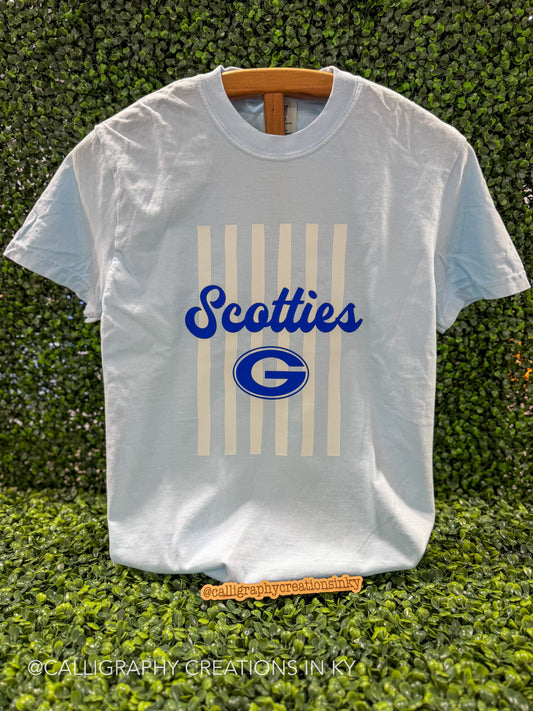 A white short-sleeve t-shirt with a blue and cream graphic print reading 'Scotties' and a large 'G' logo, displayed on a wooden hanger against a green hedge background.