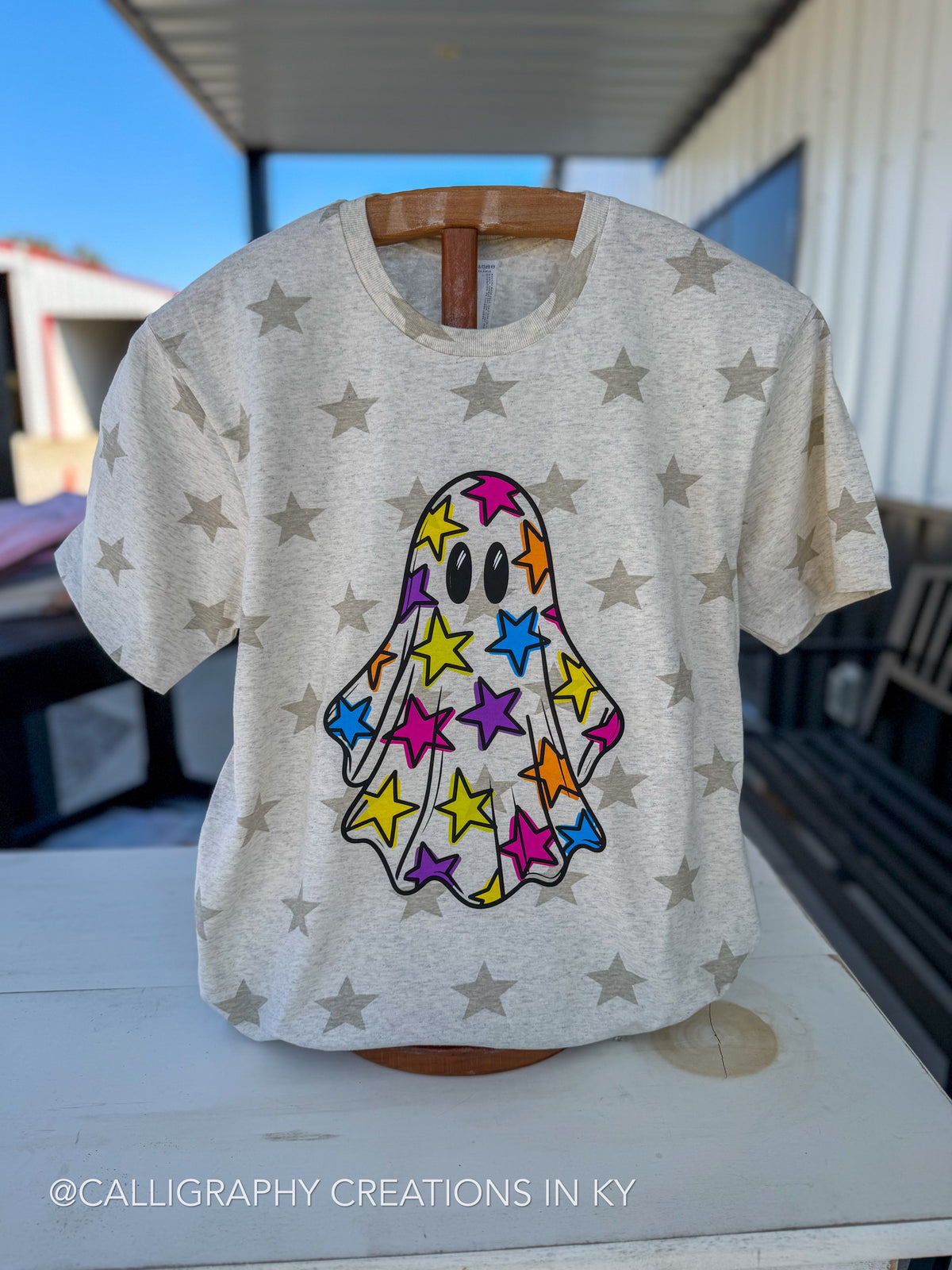 A light heather gray t-shirt with a colorful ghost made of stars on the front, displayed on a wooden hanger on an outdoor patio table.