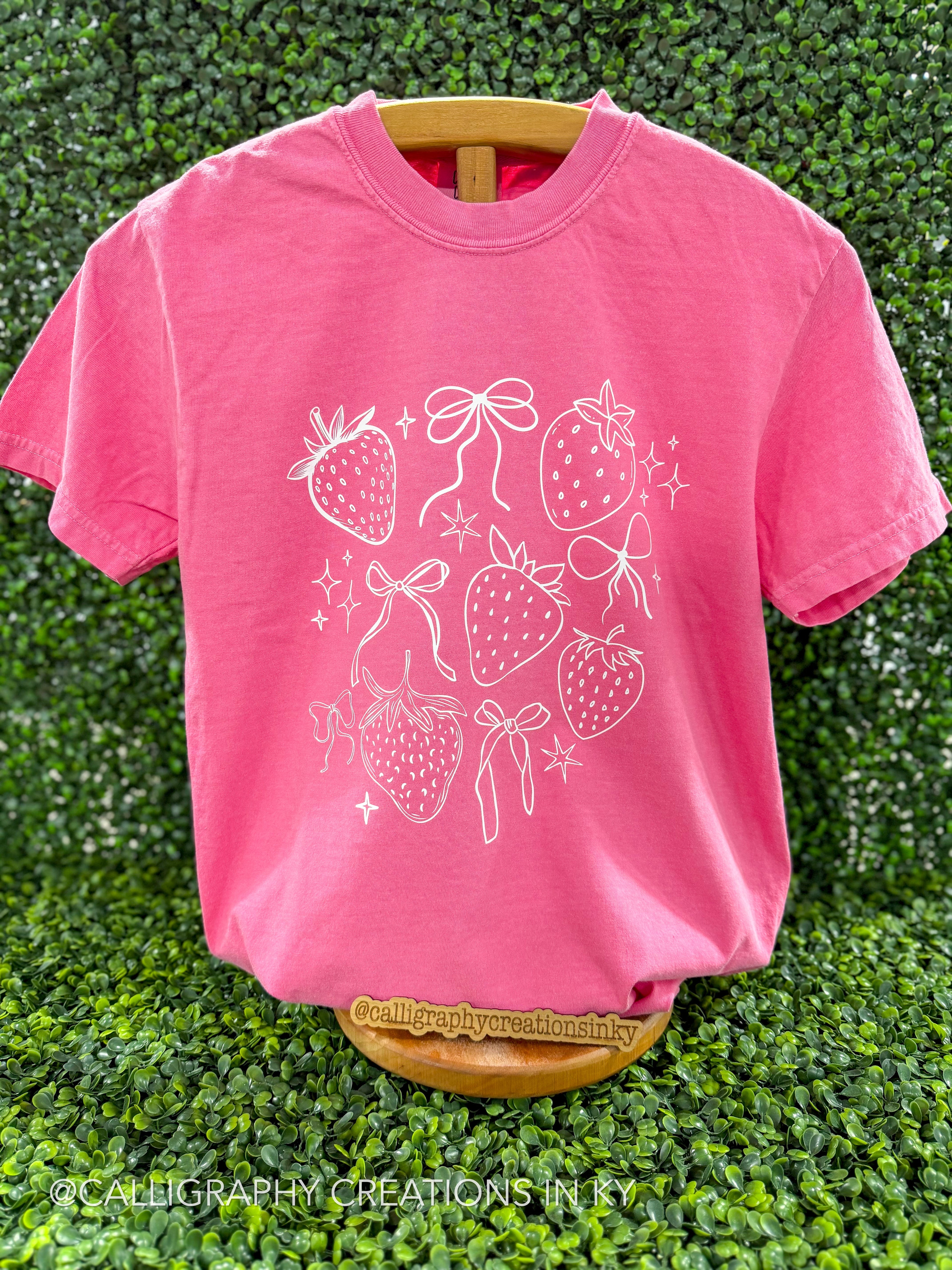 Pink shirt with strawberries and bows printed in white on a wooden stand
