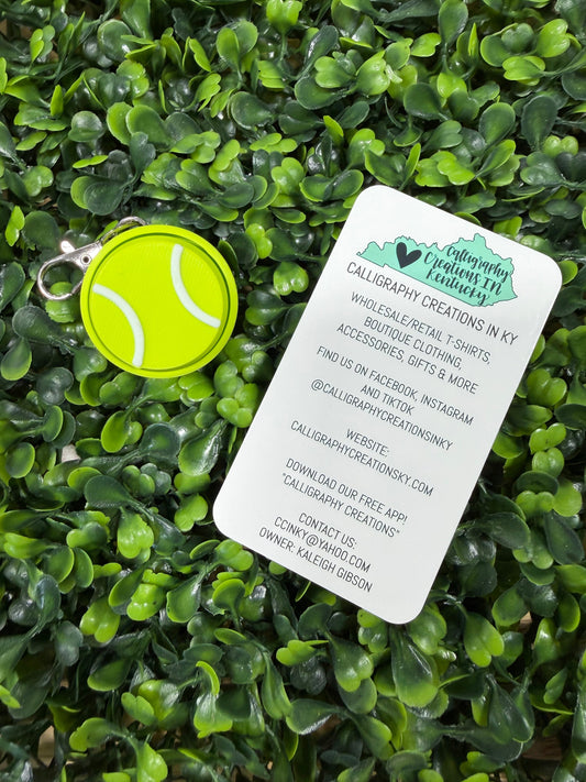 A bright yellow-green tennis ball-shaped keychain with a silver metal ring lies on a white printed business card, both placed on dense green foliage. The card shows the brand Calligraphy Creations in Ky and contact details.