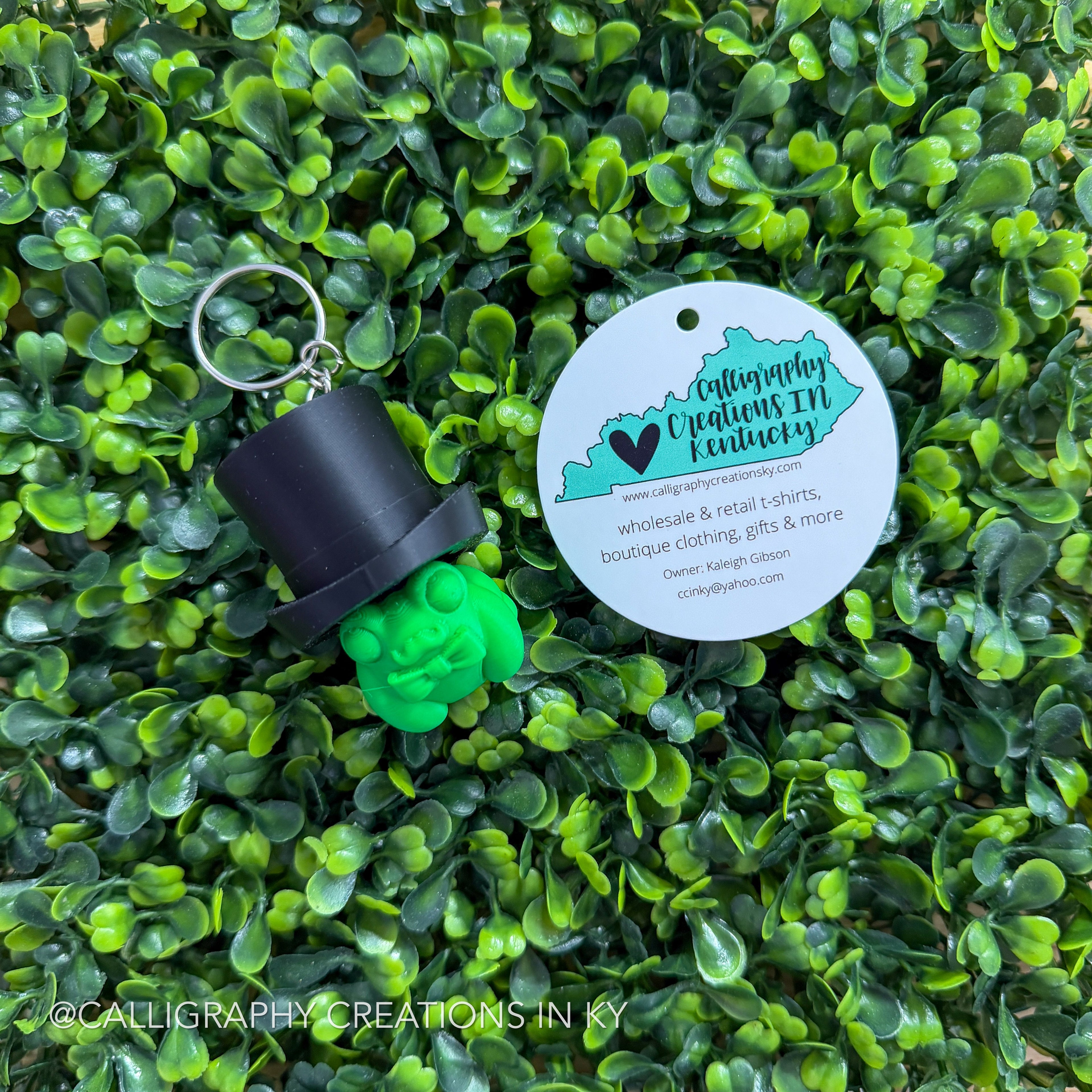 Green 3D printed frog with black top hat on greenery background with Calligraphy Creations logo.