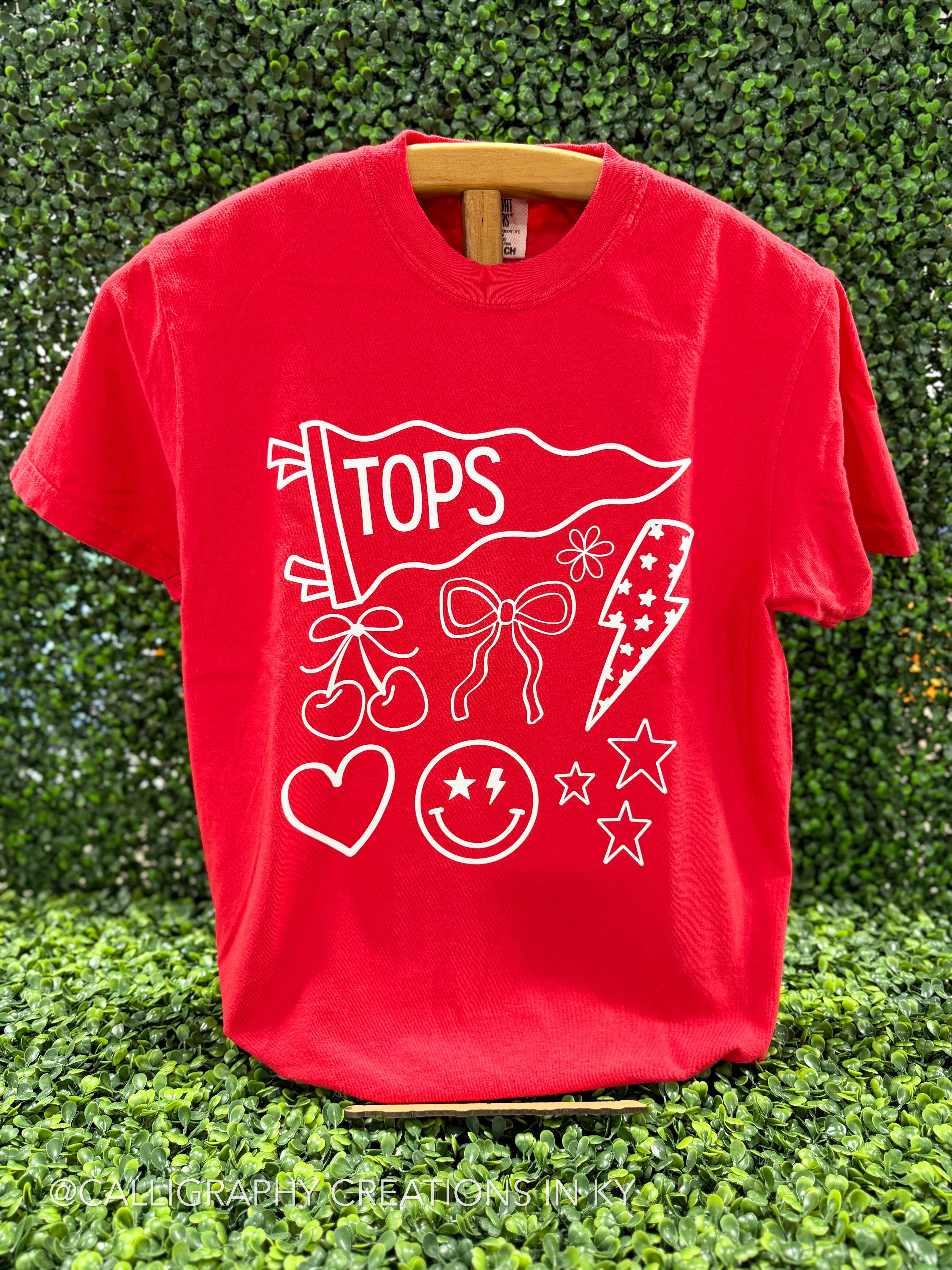 A bright red short-sleeve t-shirt with a white graphic print featuring the word 'TOPS' on a flag, bows, cherries, a lightning bolt, a smiley face, and stars, displayed on a wooden hanger against a green hedge background.