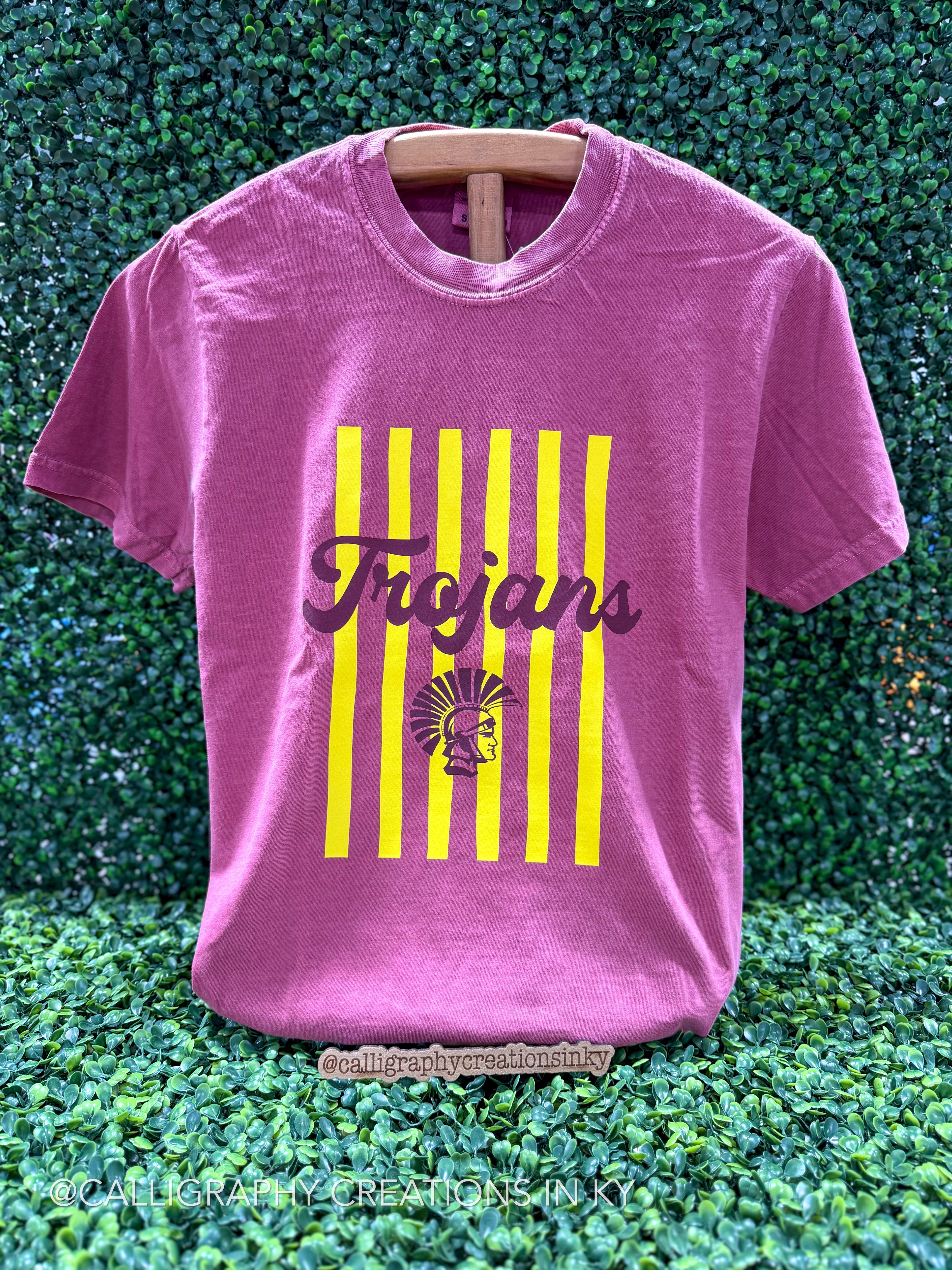 A brick short-sleeve t-shirt with a yellow/gold vertical stripe graphic and the word 'Trojans' in burgundy script, displayed on a wooden hanger against a green leaf wall background.
