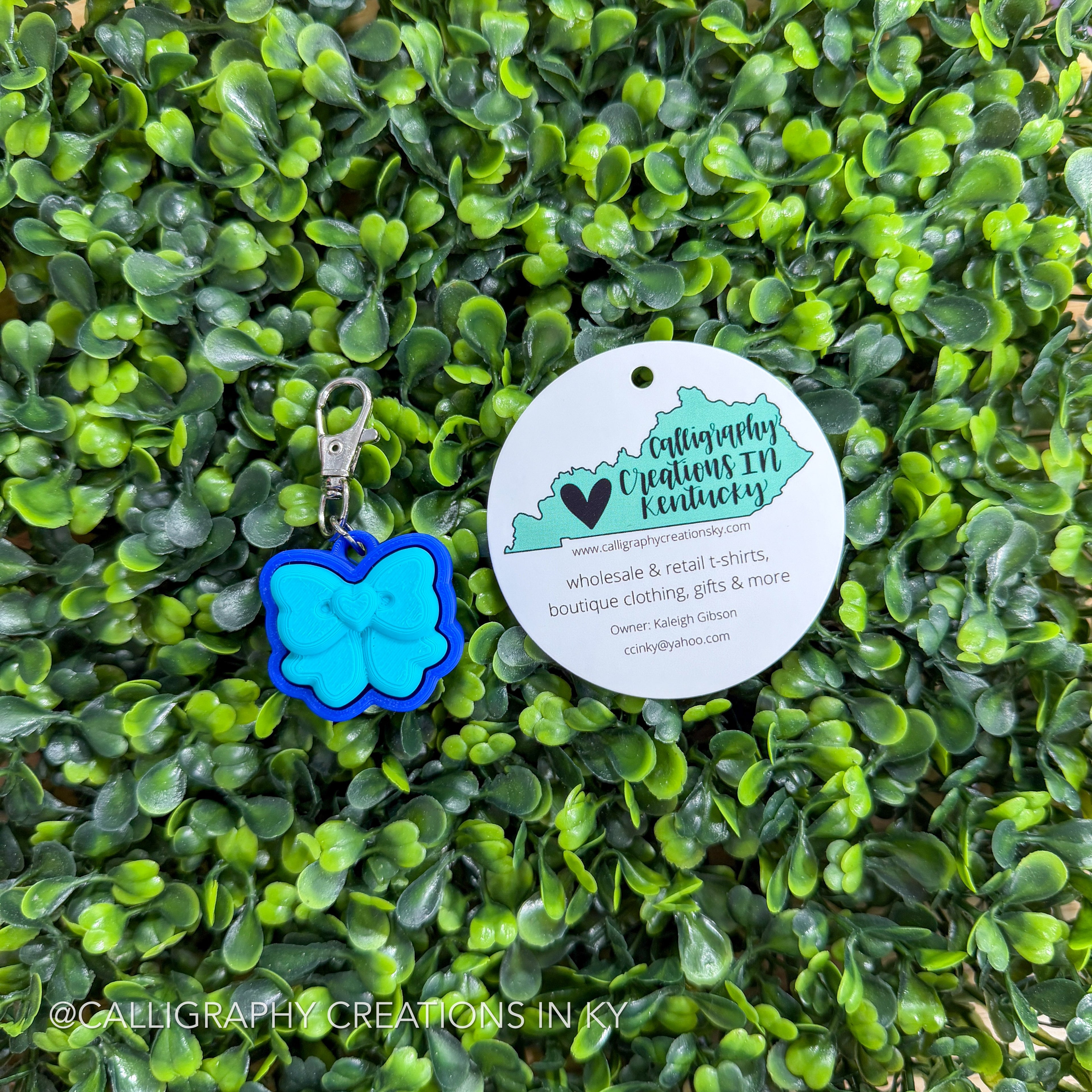 Blue bow-shaped fidget keychain with a round tag on a green leafy background