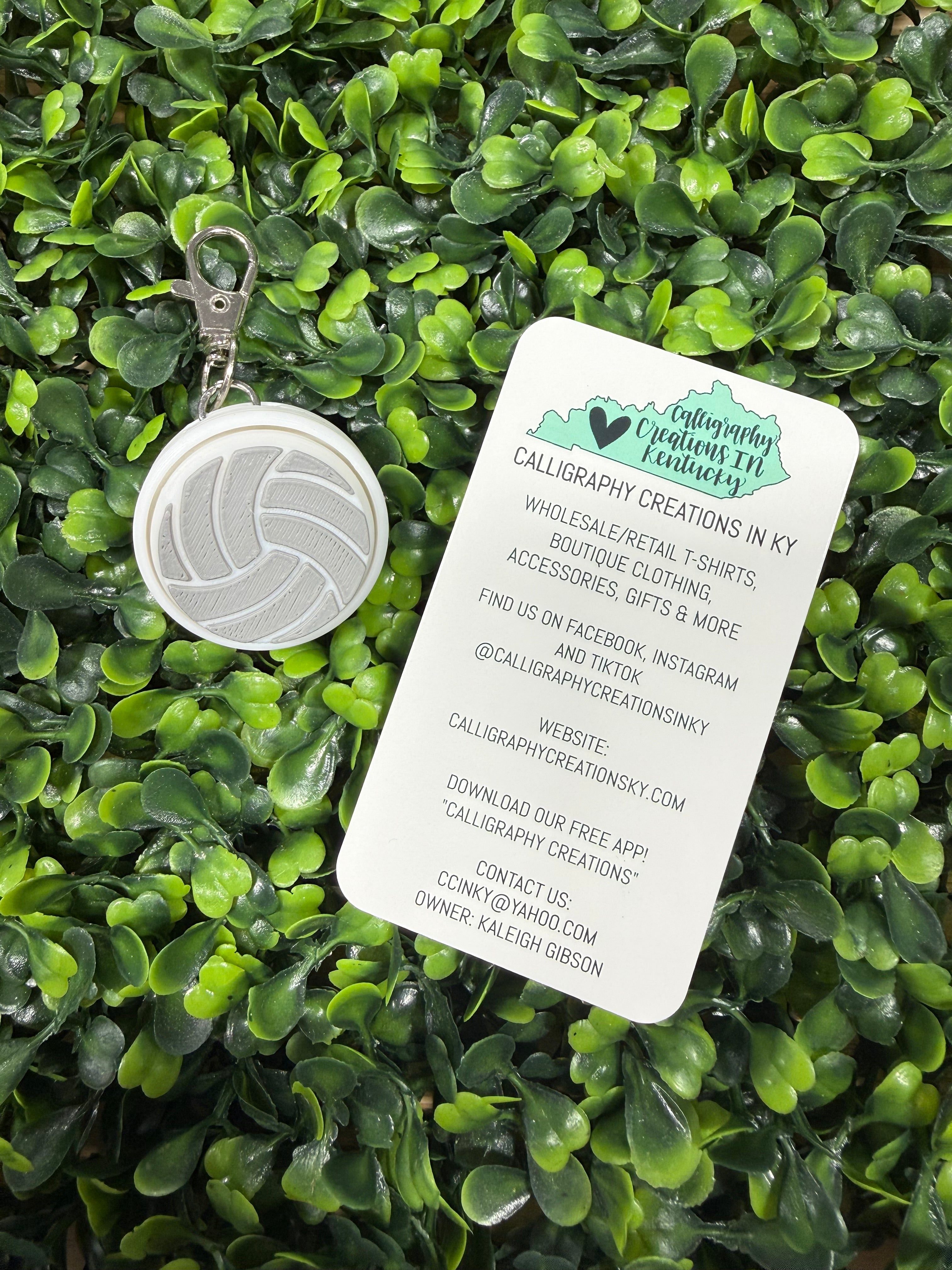 A white volleyball-themed keychain with a silver metal clasp lies on a bed of green artificial leaves, next to a white paper tag with printed business details.