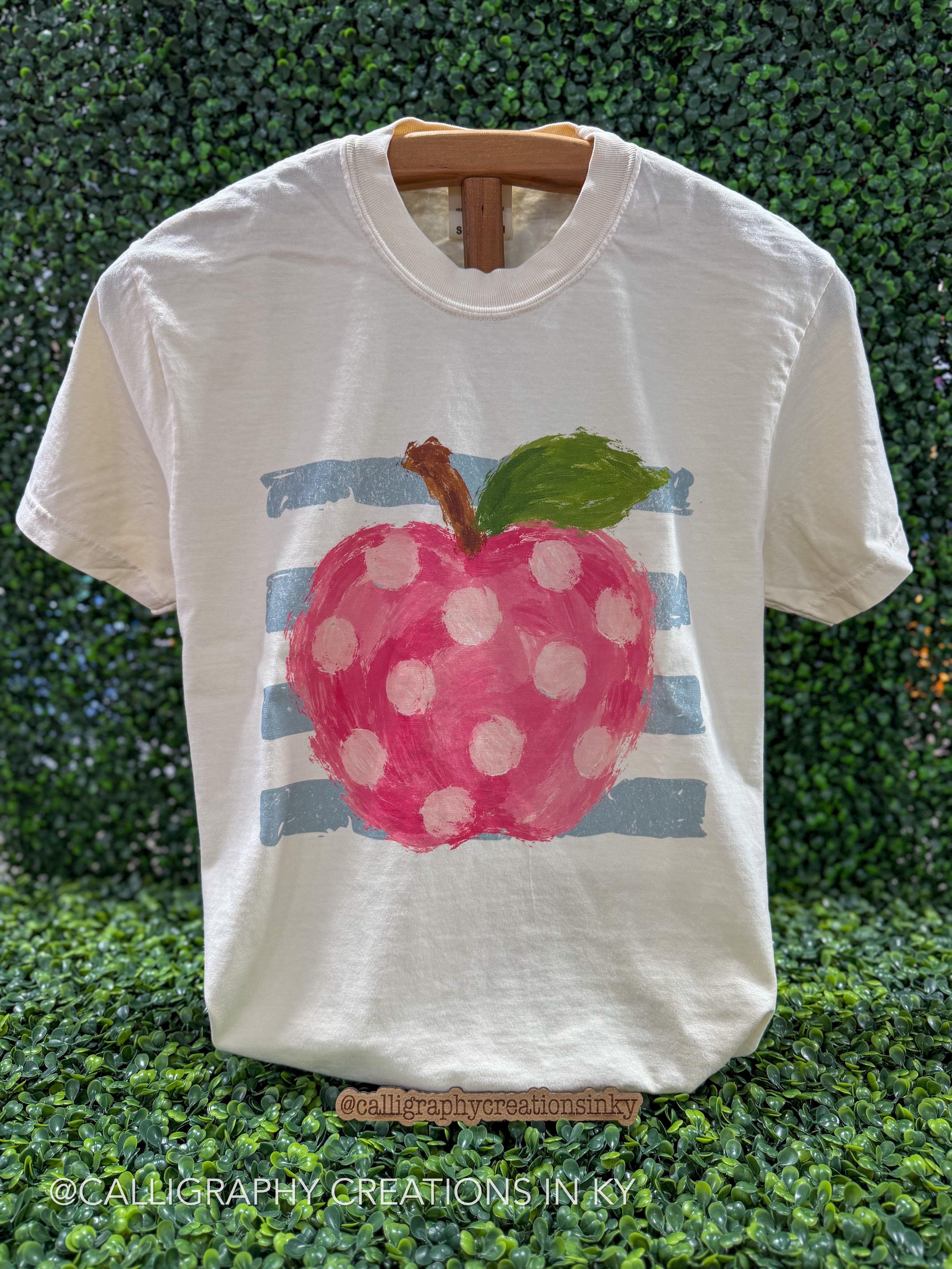 A white short-sleeve t-shirt displayed on a wooden hanger against a green leafy hedge background. The shirt features a painted graphic of a pink apple with white polka dots, a green leaf, and light blue horizontal stripes.