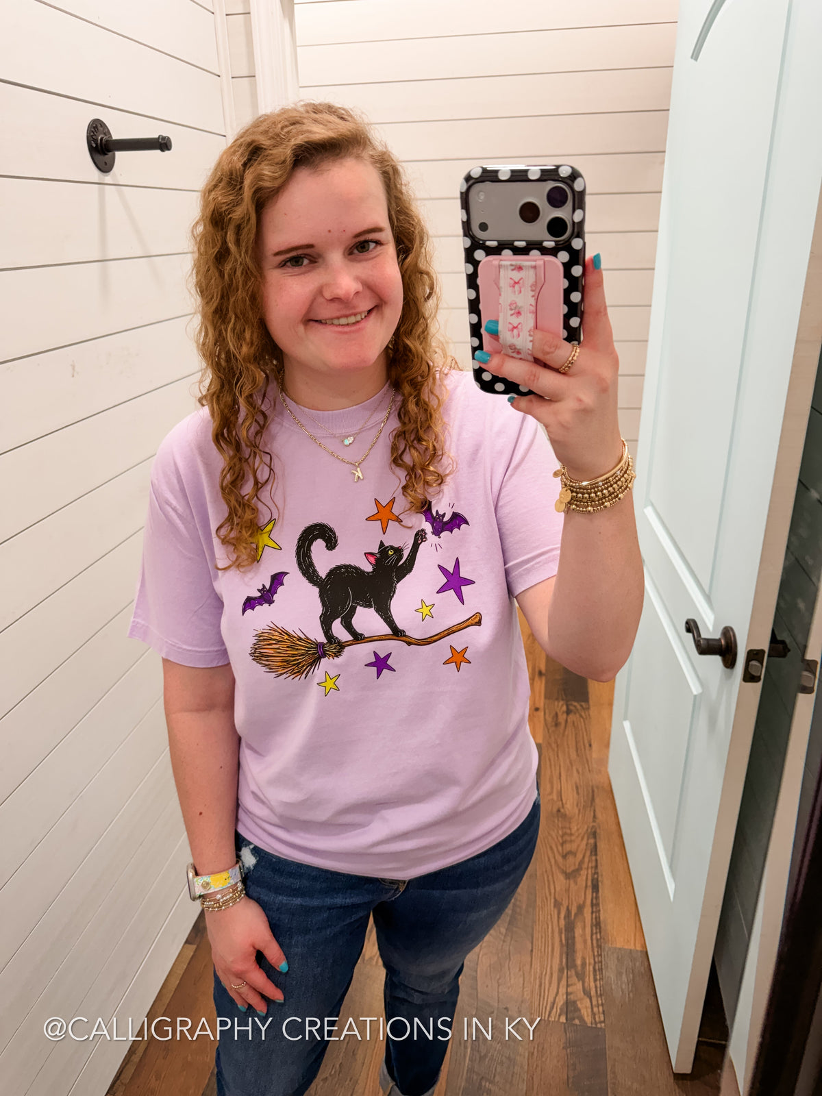 A woman takes a mirror selfie wearing a light purple t-shirt with a black witch cat graphic on a broomstick surrounded by purple and orange stars, paired with blue jeans.
