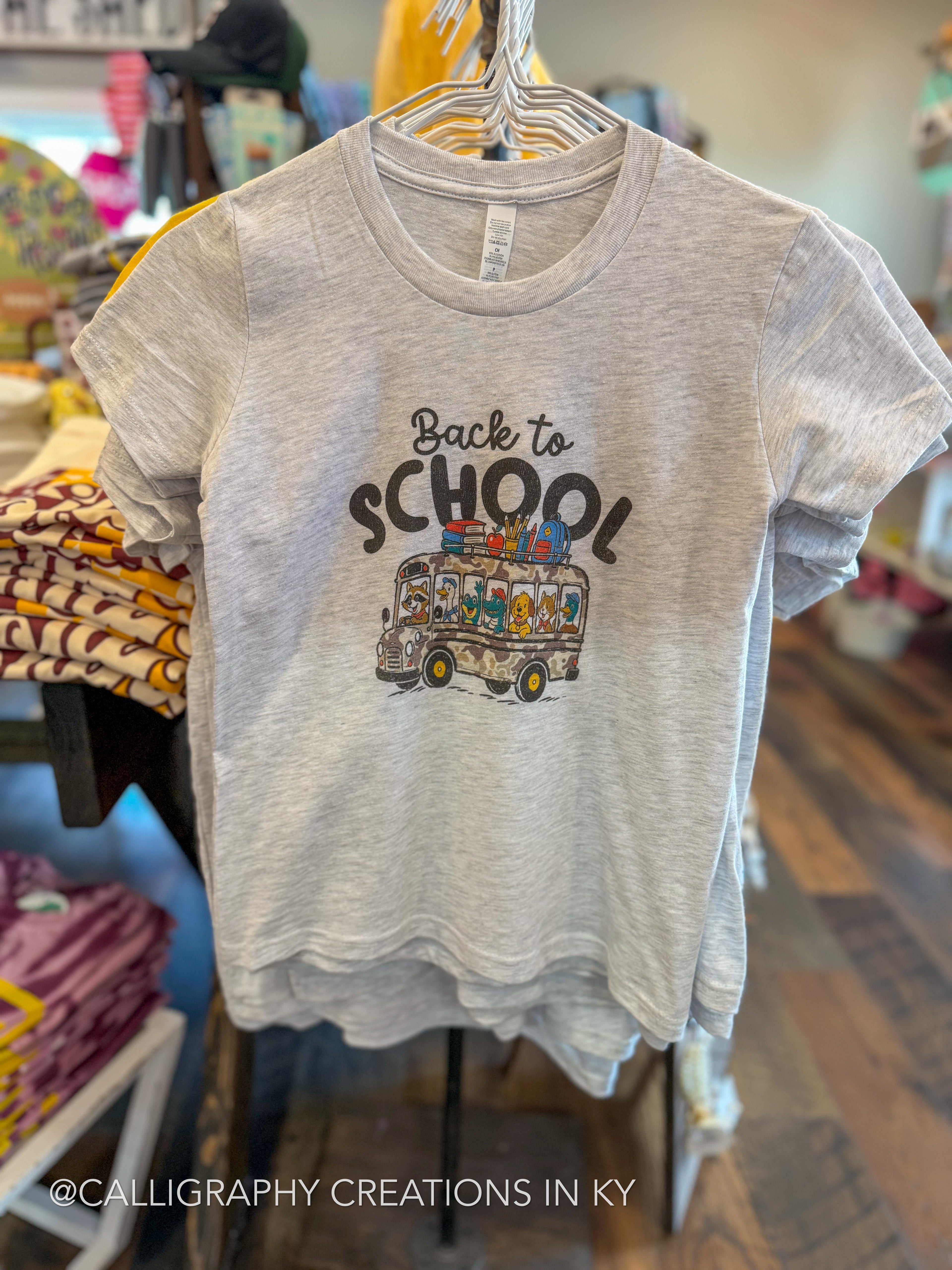 A light heather gray t-shirt hanging on a hanger in a retail store, featuring a 'Back to School' graphic with a colorful school bus and black text.