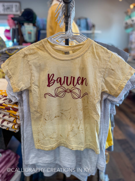 A short-sleeve graphic t-shirt in a warm, faded yellow-beige color, hanging on a hanger in a retail store. The shirt features the word 'Barren' in dark burgundy cursive script with a matching ribbon/bow illustration underneath.