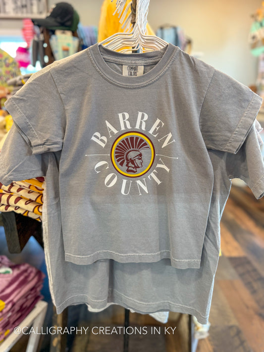 A gray short-sleeve t-shirt hanging on a hanger in a retail store, featuring a distressed graphic on the front that reads 'BARREN COUNTY' in white around a circular emblem with a Trojan head in yellow, red, and brown.