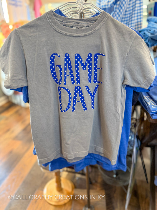 A heather gray short-sleeve t-shirt hanging on a metal hanger, featuring the words 'GAME DAY' in blue letters with white polka dots. The shirt is layered over a blue t-shirt in a retail store setting.