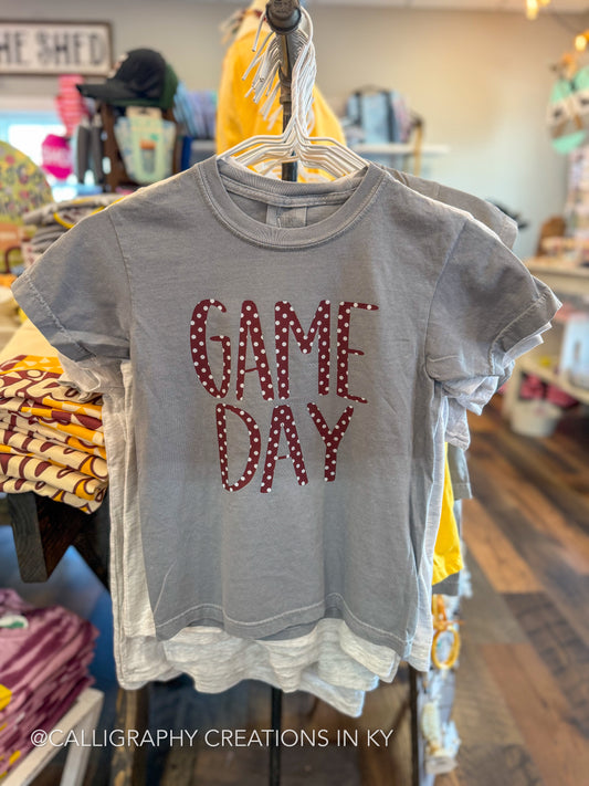 A gray short-sleeve t-shirt with 'GAME DAY' in burgundy letters filled with white polka dots, hanging on a hanger inside a retail store.