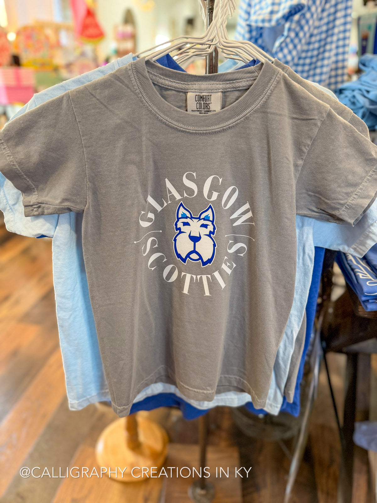 A gray short-sleeve graphic T-shirt hanging on a hanger in a retail store. The shirt features a circular white and blue 'GLASGOW SCOTTIES' design with a dog mascot logo in the center.
