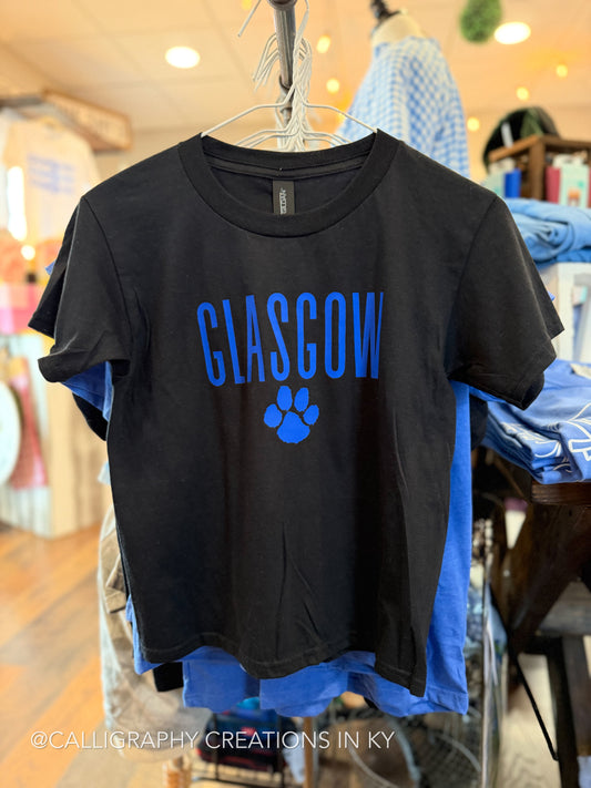 A black short-sleeve T-shirt hanging on a hanger in a retail store, featuring the word 'GLASGOW' in blue block letters and a blue paw print graphic on the front.
