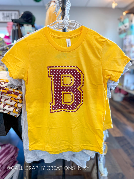 A bright yellow short-sleeve t-shirt hanging on a hanger in a retail store. The shirt features a large decorative letter 'B' on the front, outlined in gold and filled with dark burgundy polka dots.