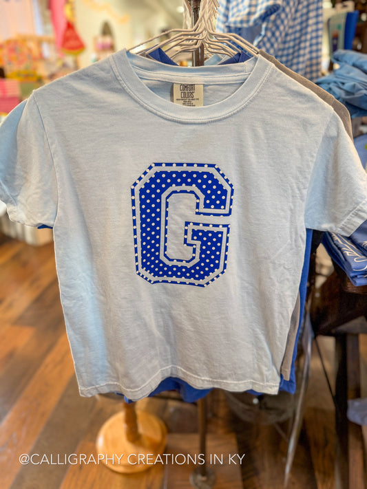 A light blue short-sleeve crop top t-shirt hanging on a metal hanger, featuring a large blue and white polka-dot letter 'G' graphic on the front. The shirt is displayed in a retail store setting with wooden floors and other clothing visible in the background.