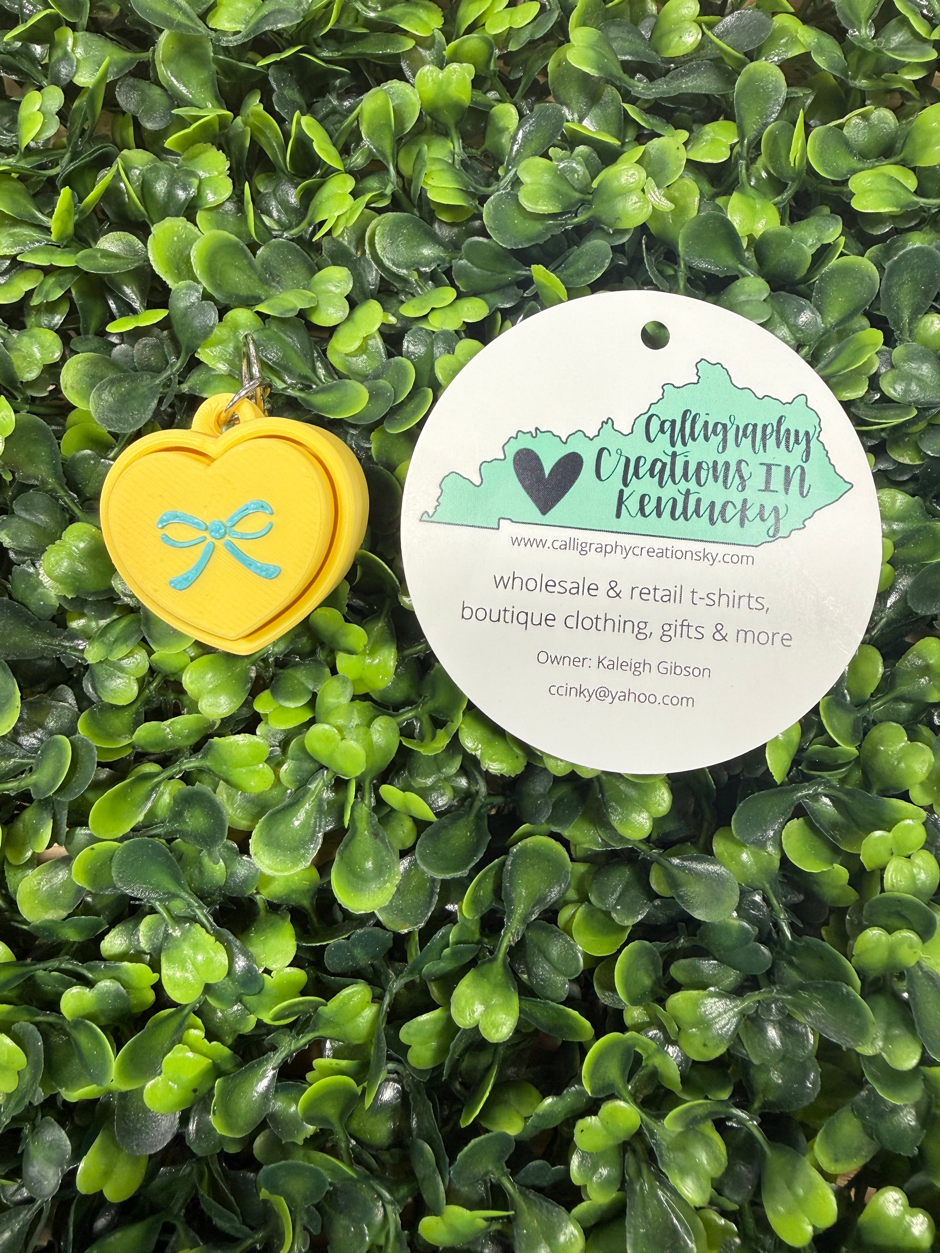 Yellow heart key chain with teal bow on a bed of green leaves