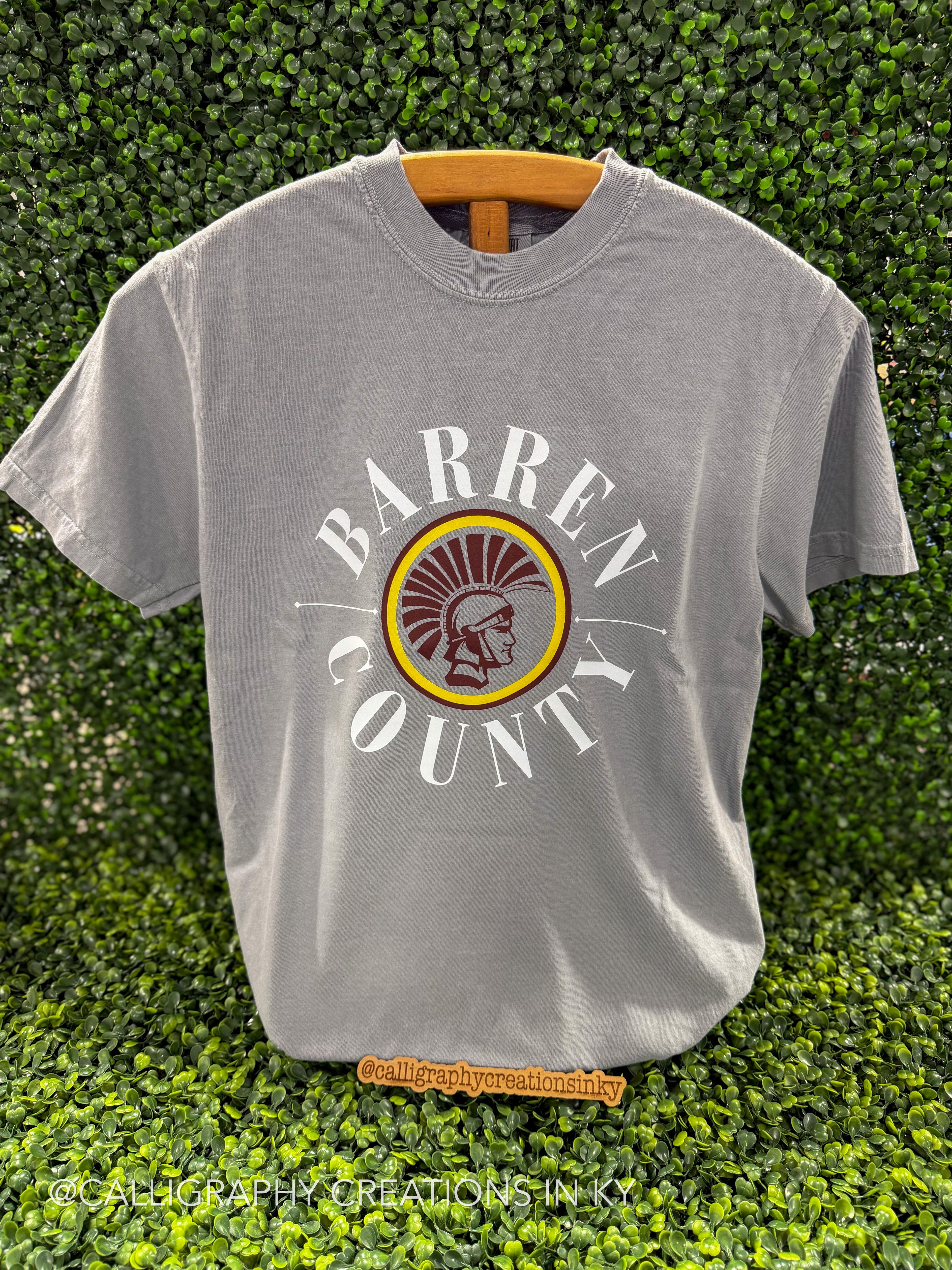 A heather gray short-sleeve t-shirt hanging on a wooden hanger against a green leafy hedge background. The shirt features a circular graphic in the center with the words 'BARREN COUNTY' in white arched around a circular emblem containing a brown and yellow radiating sunburst and a white profile head.