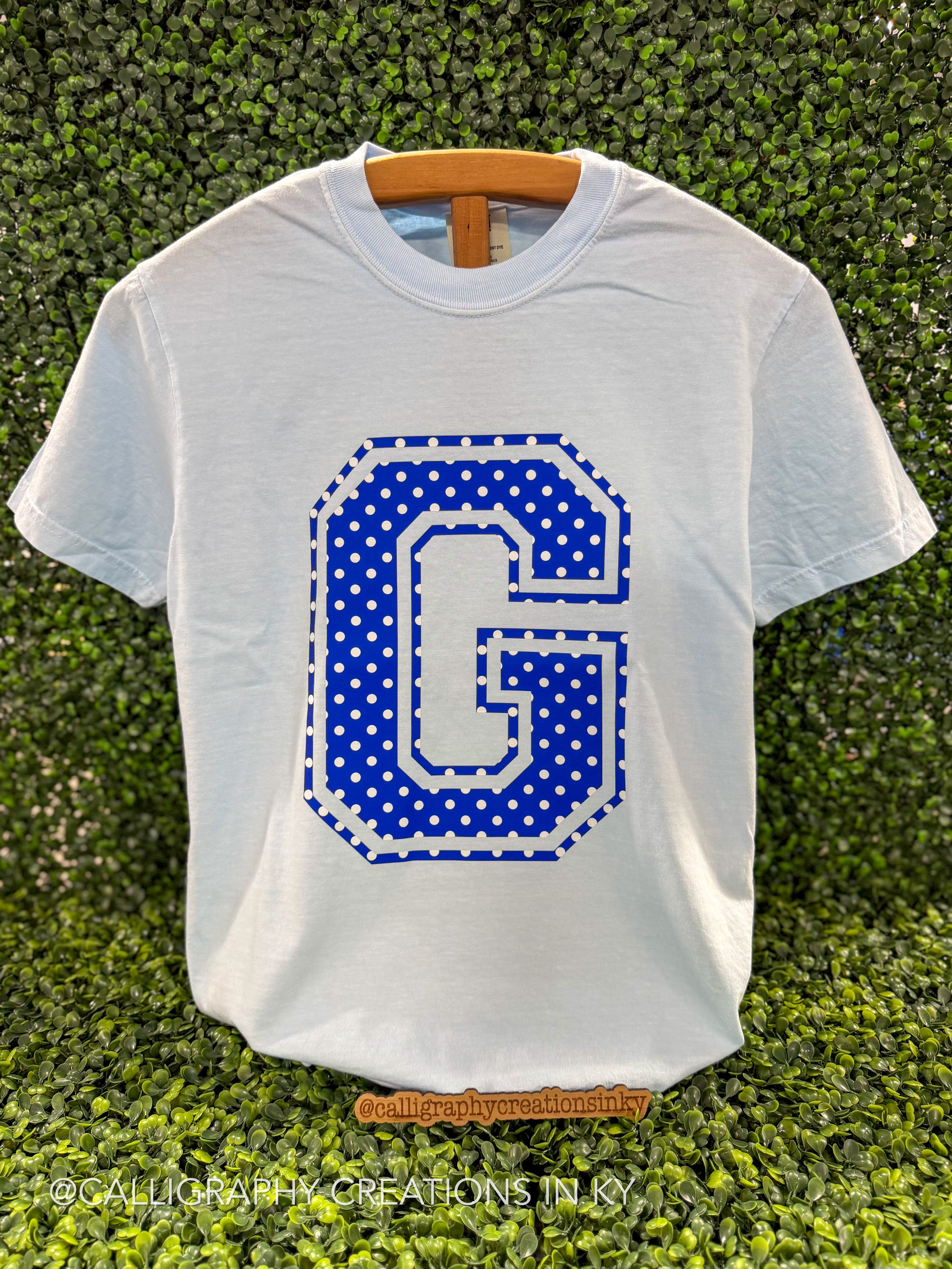 A white short-sleeve t-shirt with a large blue and white polka-dot letter 'G' graphic on the front, displayed on a wooden hanger against a green hedge background.