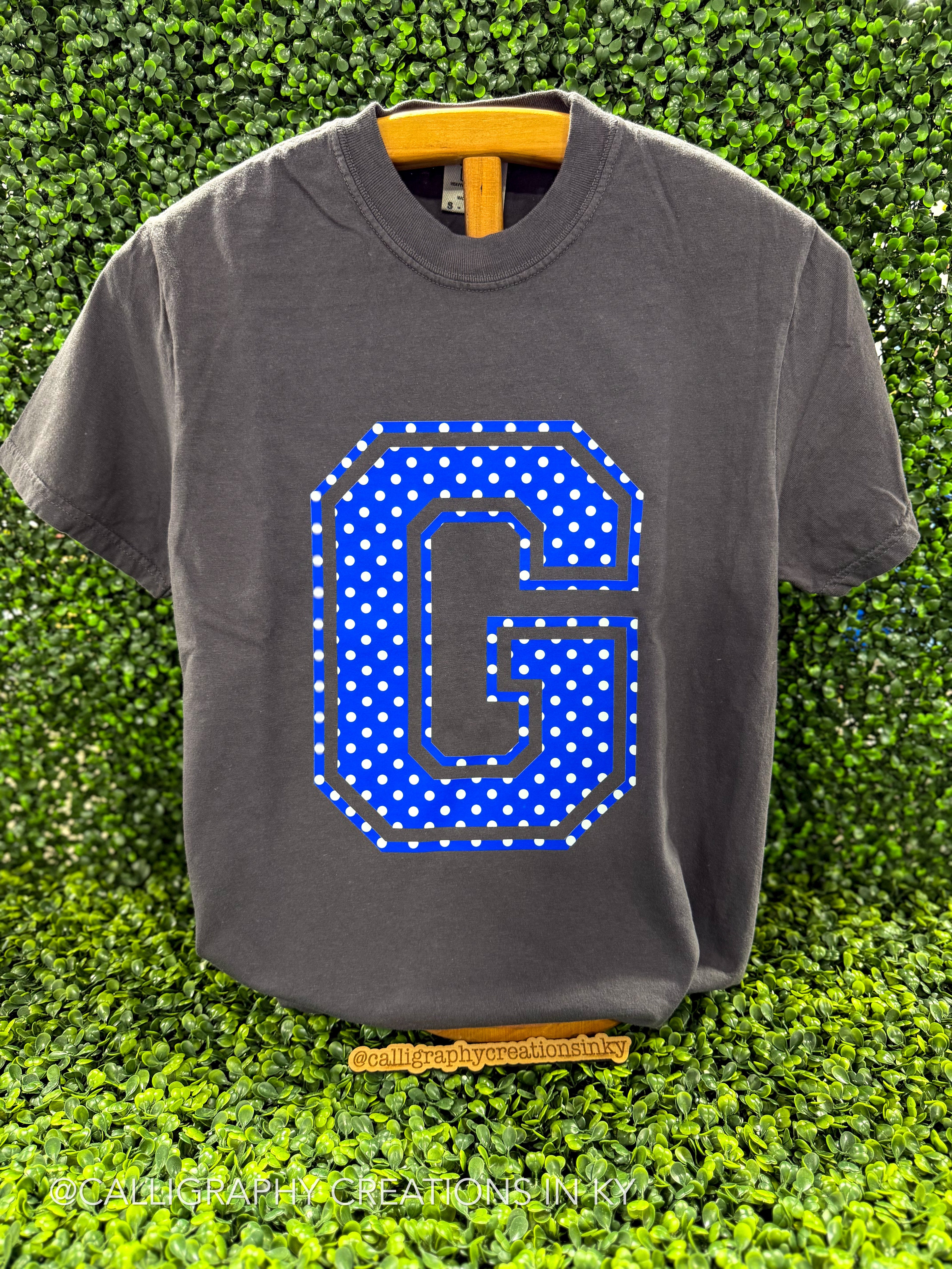 A dark gray t-shirt with a large blue and white polka dot letter 'G' graphic on the front, displayed on a wooden hanger against a green artificial hedge background.