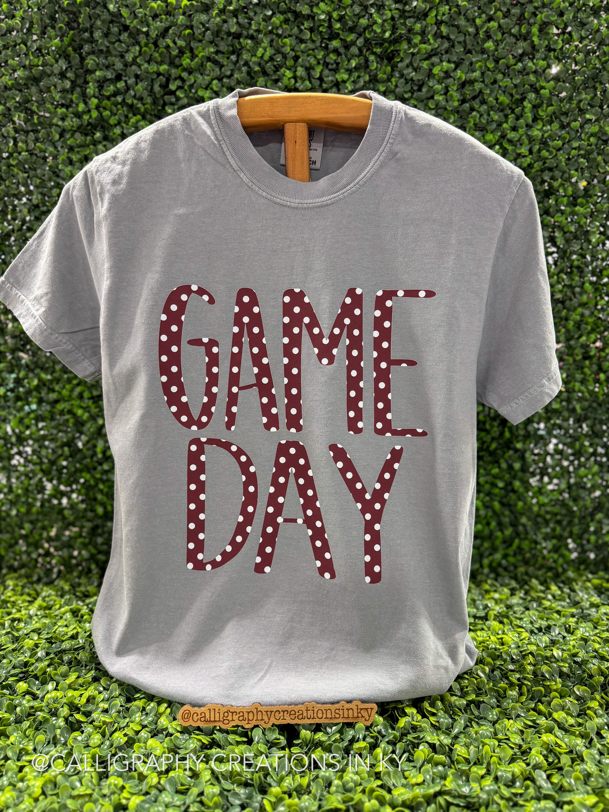 A light gray t-shirt with a wooden hanger, featuring the words 'GAME DAY' in a red and white polka dot pattern. The shirt is displayed against a green leafy hedge background.