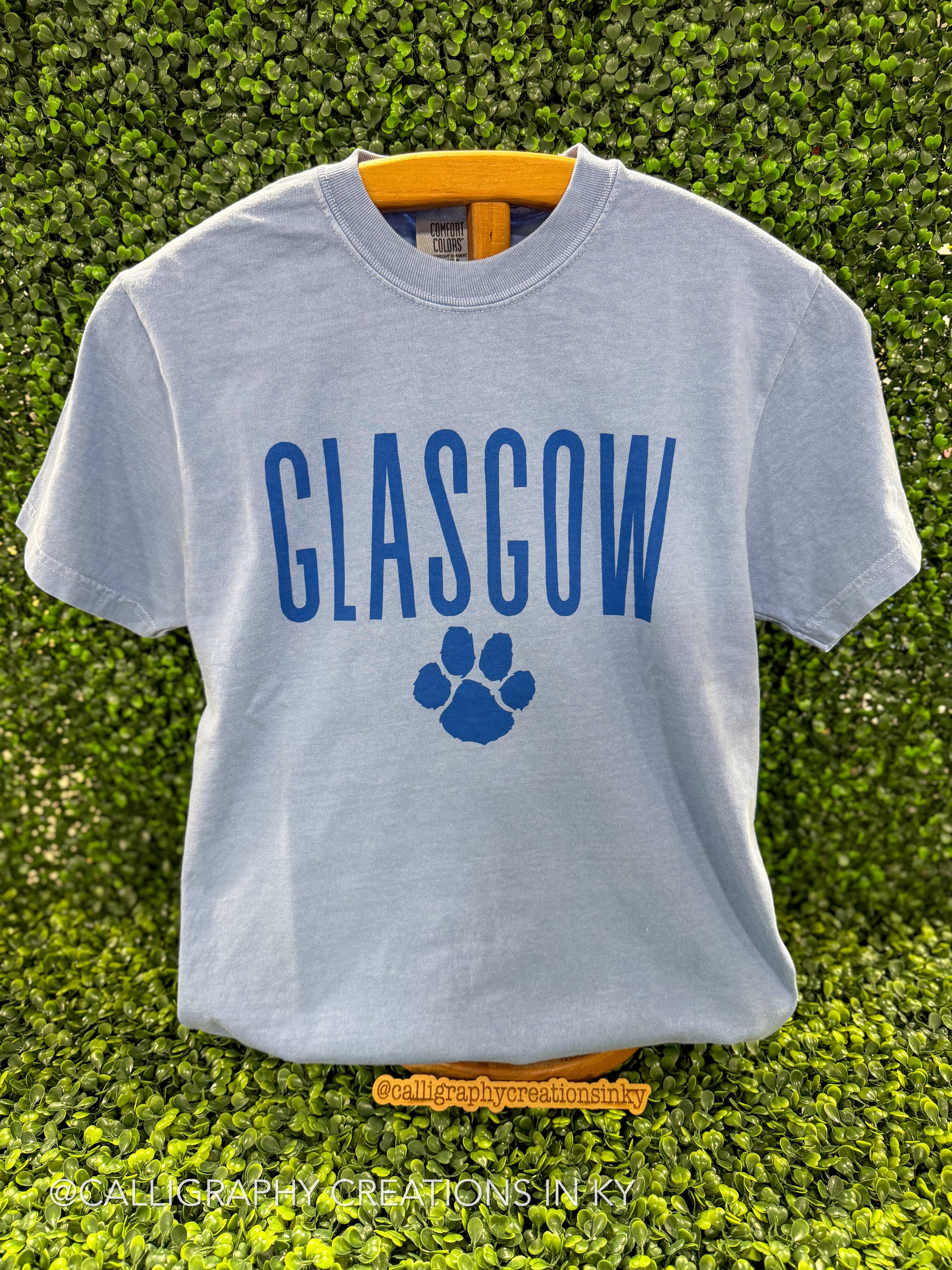 A light blue short-sleeve t-shirt with 'GLASGOW' and a paw print graphic in dark blue, displayed on a yellow hanger against a green leaf wall background.