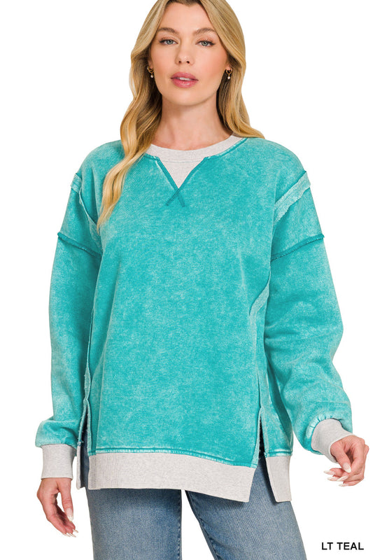 A woman modeling a teal acid-wash long-sleeve sweatshirt with a gray ribbed crew neck, cuffs, and hem, paired with blue jeans against a white background.