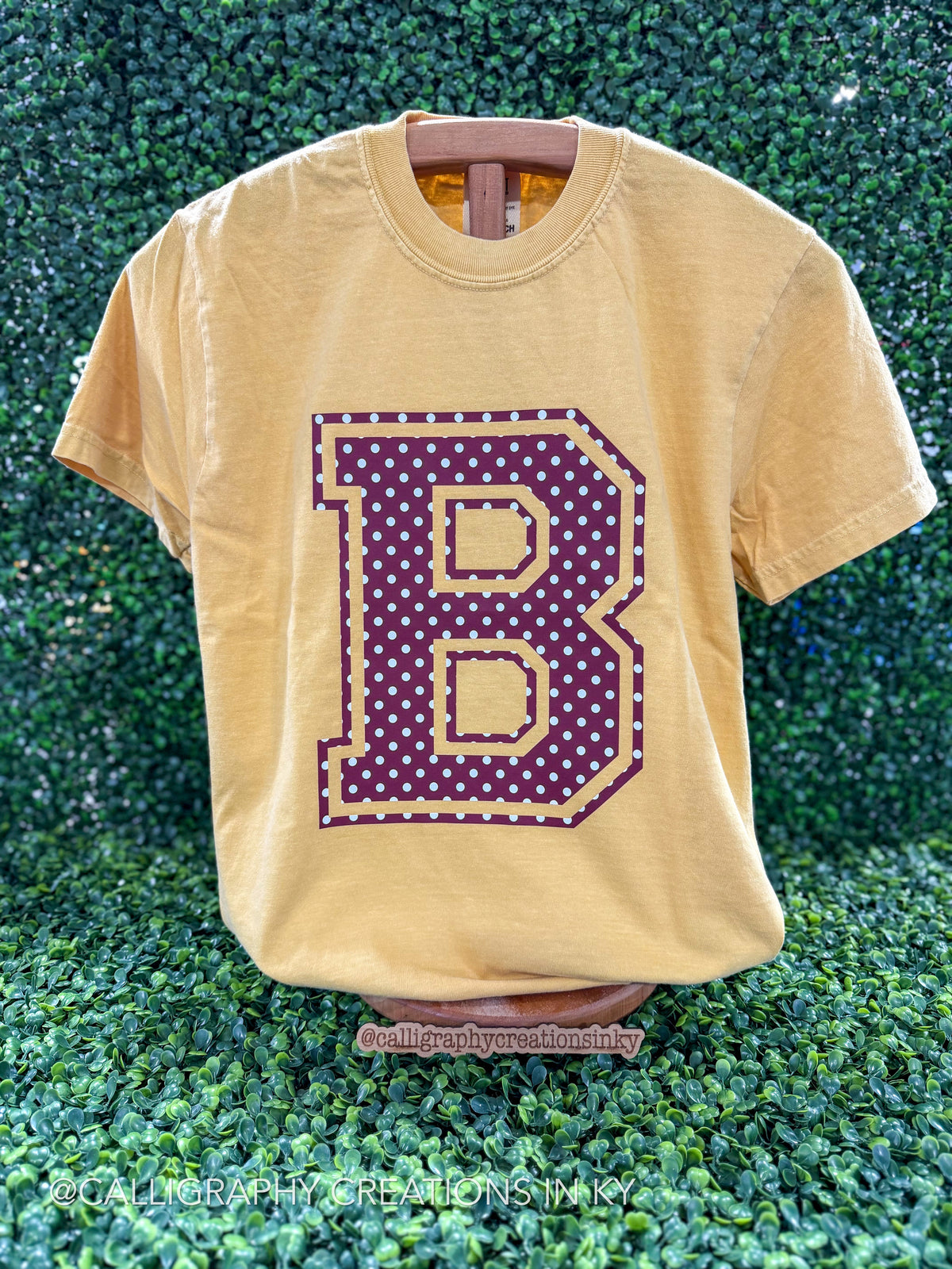A faded mustard yellow t-shirt displayed on a wooden hanger against a green leafy hedge background. The shirt features a large collegiate-style letter 'B' graphic on the front, filled with purple polka dots and outlined in gold.