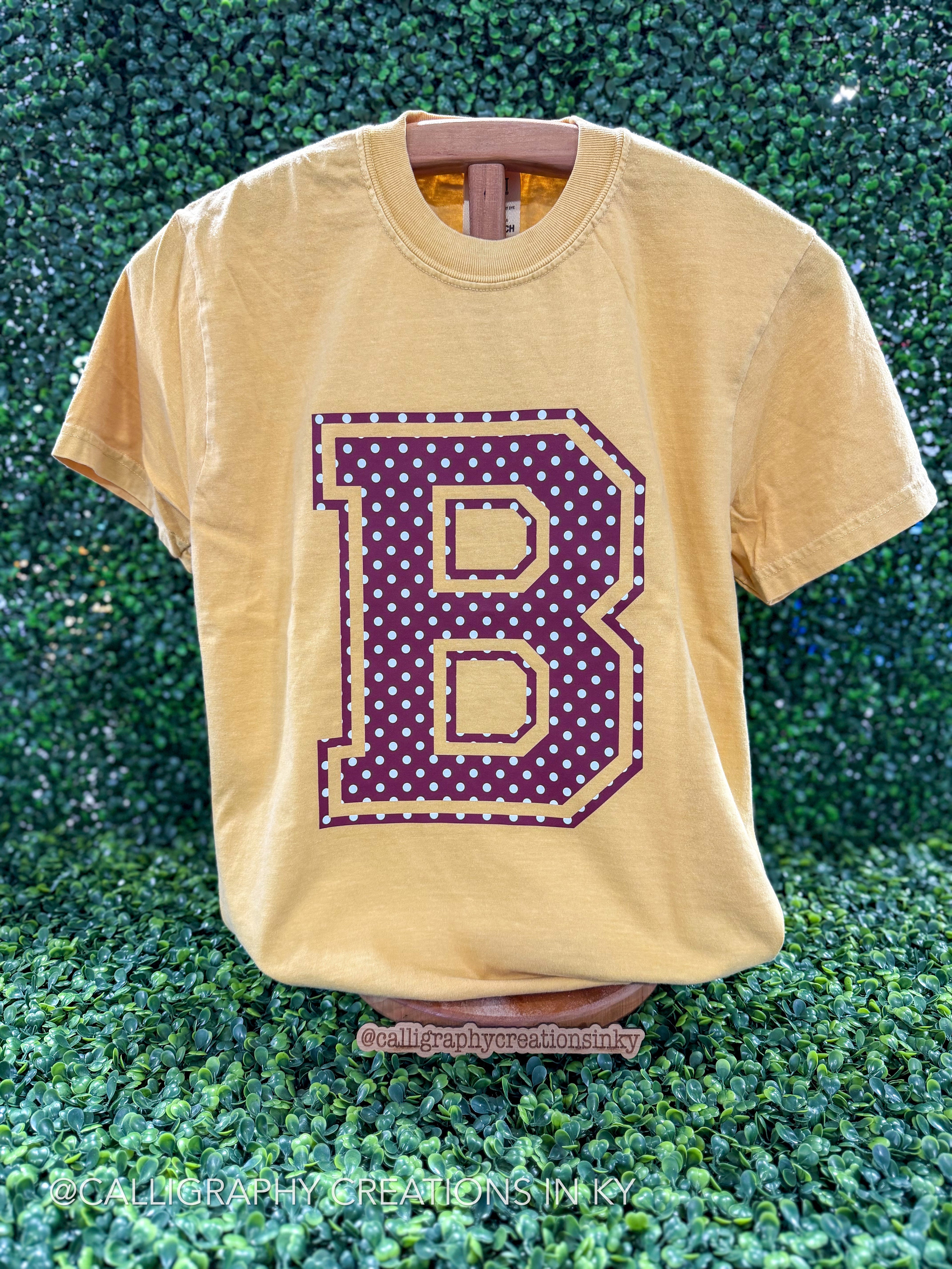 A faded mustard yellow t-shirt displayed on a wooden hanger against a green leafy hedge background. The shirt features a large collegiate-style letter 'B' graphic on the front, filled with purple polka dots and outlined in gold.