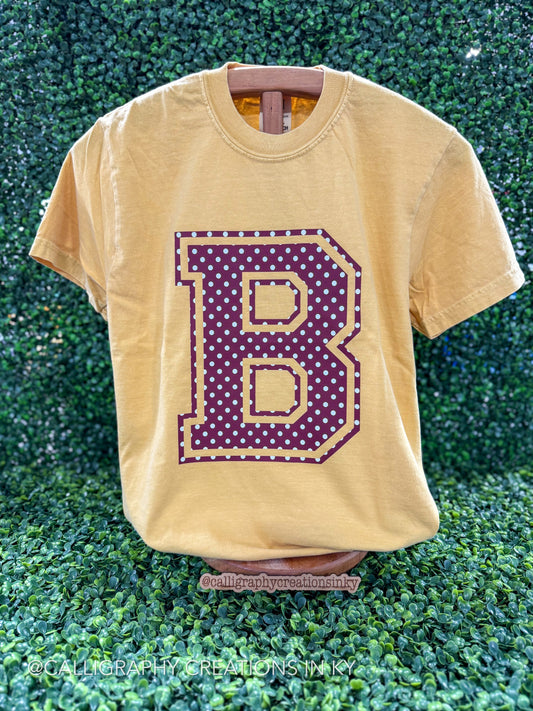A faded mustard yellow t-shirt displayed on a wooden hanger against a green leafy hedge background. The shirt features a large collegiate-style letter 'B' graphic on the front, filled with purple polka dots and outlined in gold.