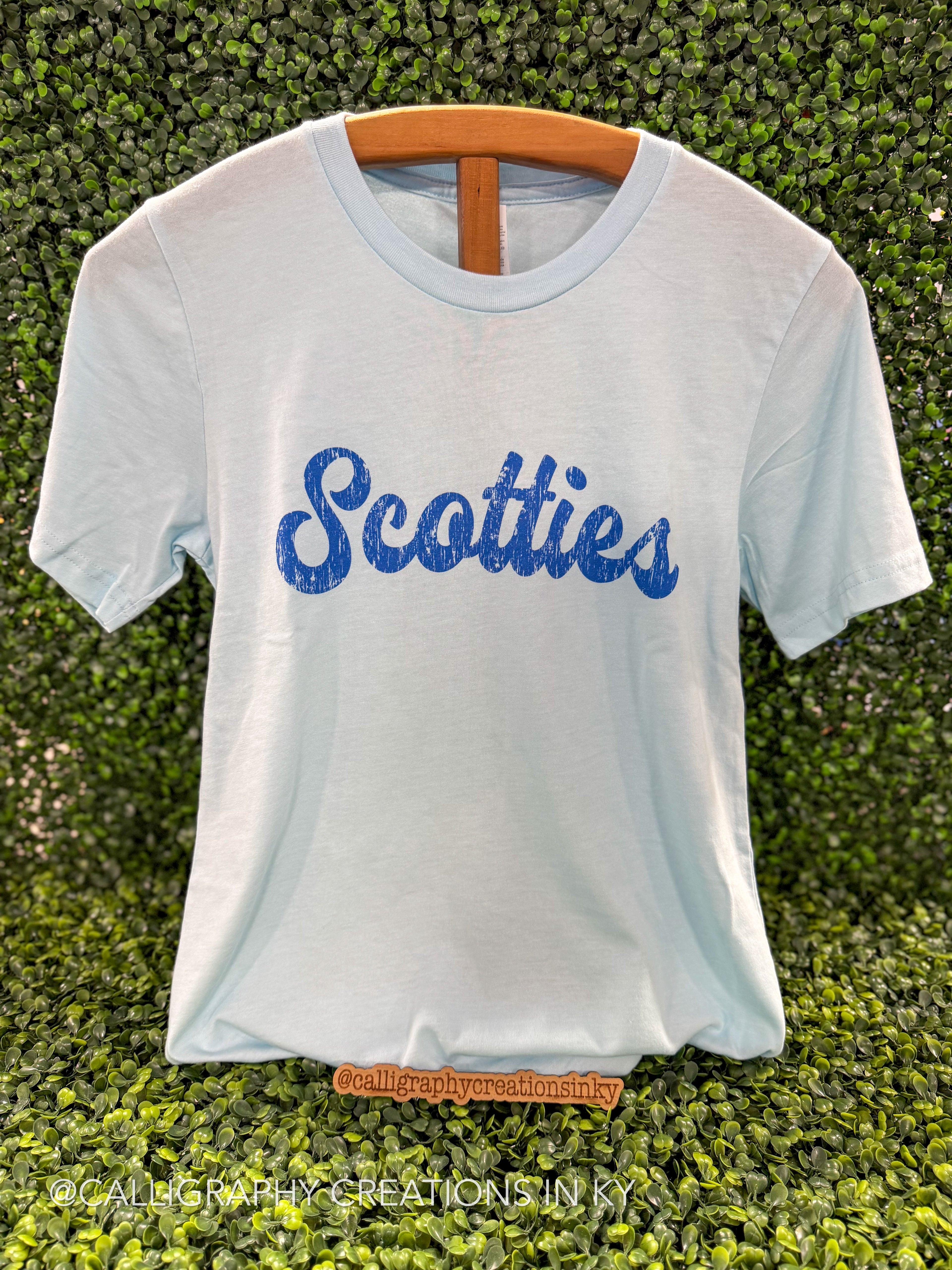 A light blue short-sleeve t-shirt hanging on a wooden hanger against a green leaf wall. The shirt features the word 'Scotties' printed across the chest in a textured blue cursive font.