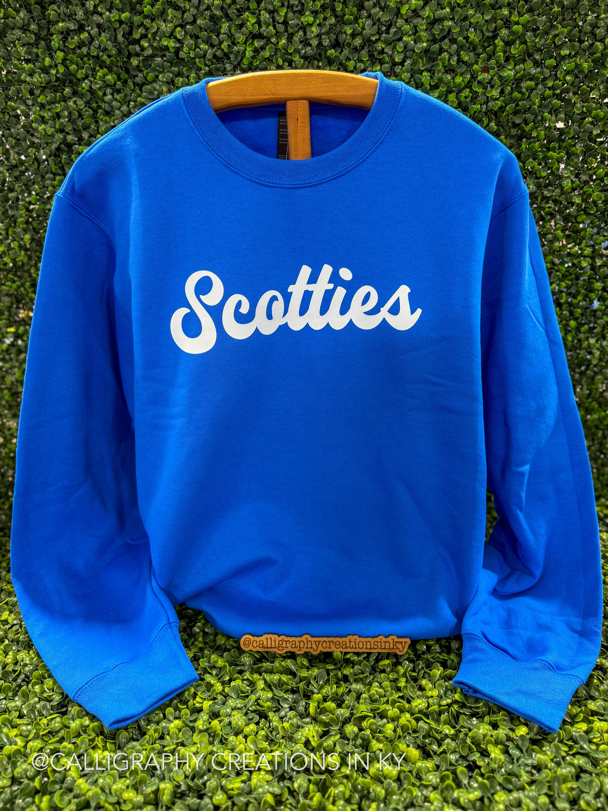 A bright blue crewneck sweatshirt with the word 'Scotties' printed in white script across the chest, displayed on a wooden hanger against a green hedge background.