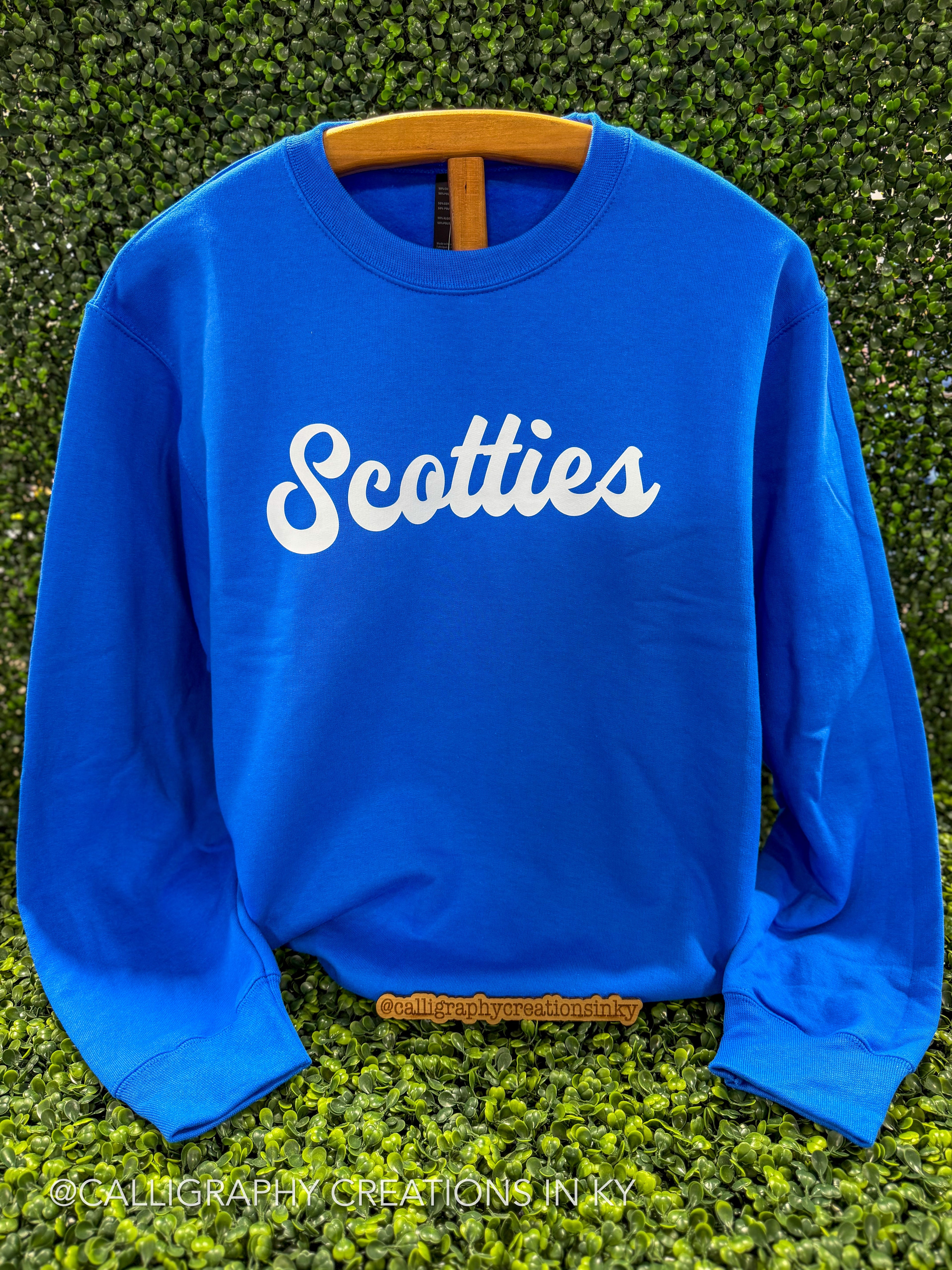 A bright blue crewneck sweatshirt with the word 'Scotties' printed in white script across the chest, displayed on a wooden hanger against a green hedge background.