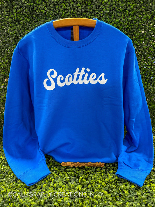 A bright blue crewneck sweatshirt with the word 'Scotties' printed in white script across the chest, displayed on a wooden hanger against a green hedge background.