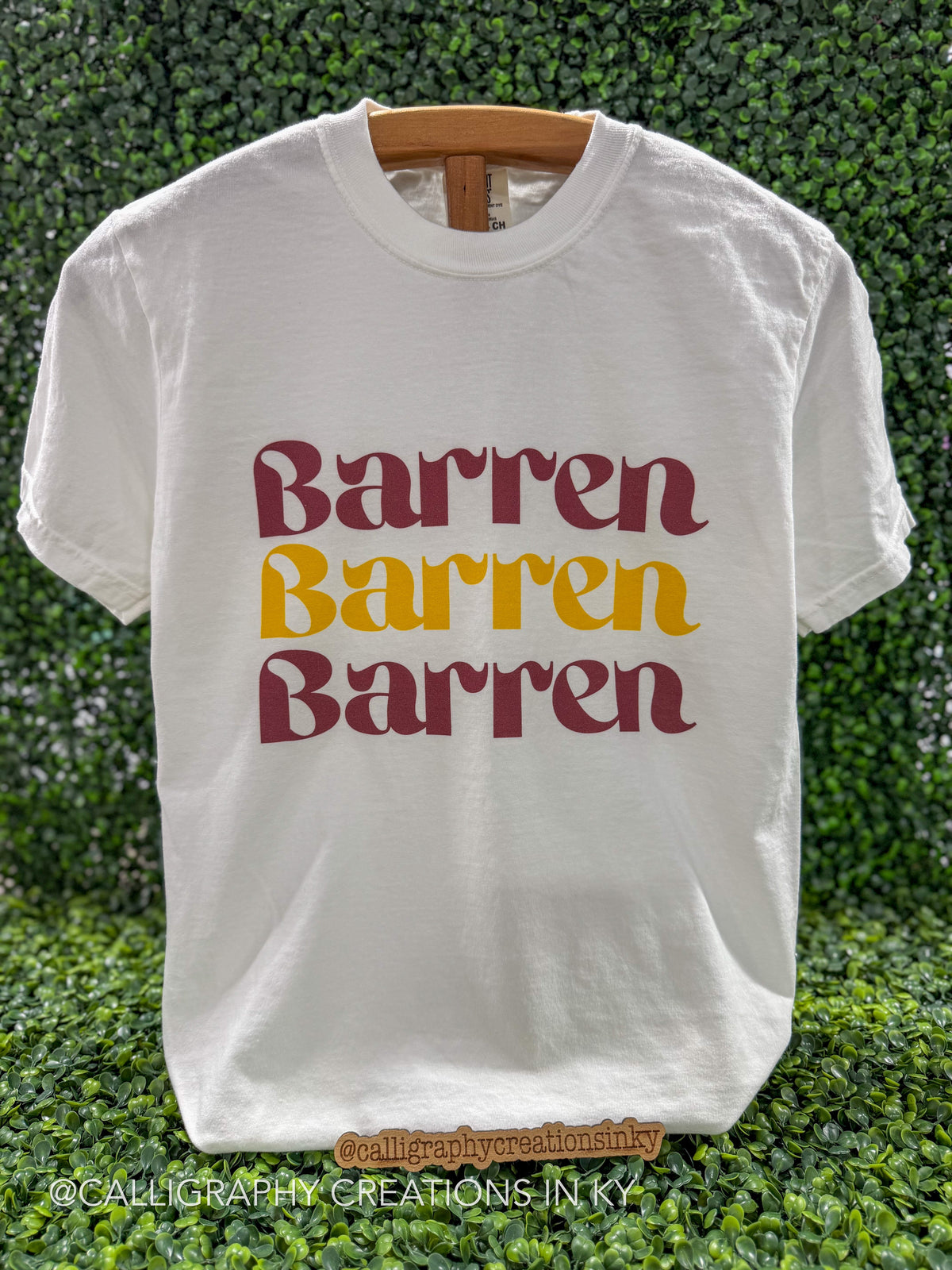 A white short-sleeve t-shirt with the word 'Barren' printed three times in a wavy font, alternating between dark red and mustard yellow. The shirt is displayed on a wooden hanger against a green leafy hedge background.