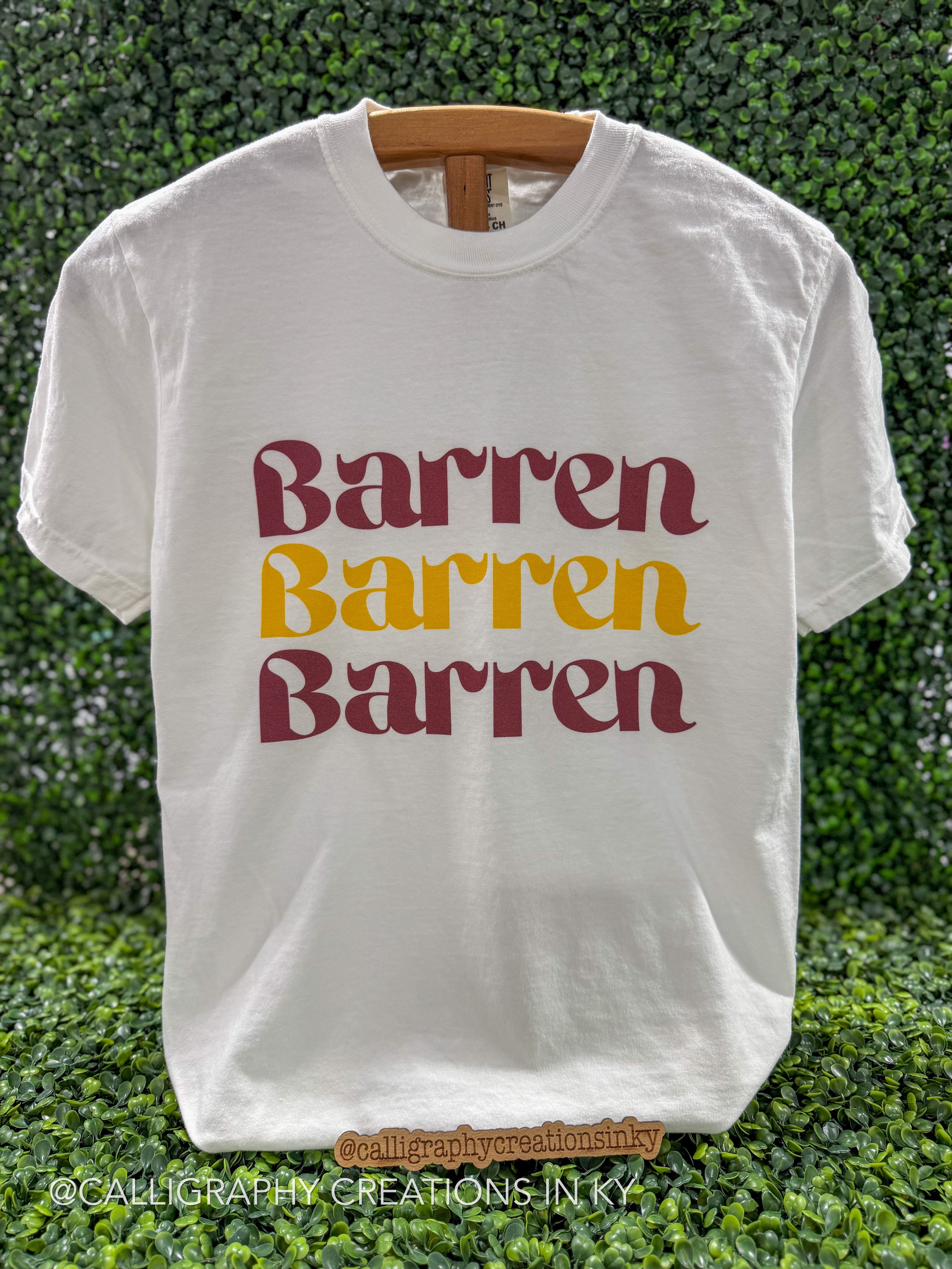 A white short-sleeve t-shirt with the word 'Barren' printed three times in a wavy font, alternating between dark red and mustard yellow. The shirt is displayed on a wooden hanger against a green leafy hedge background.