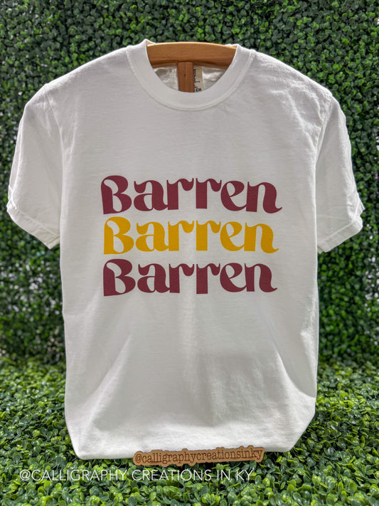 A white short-sleeve t-shirt with the word 'Barren' printed three times in a wavy font, alternating between dark red and mustard yellow. The shirt is displayed on a wooden hanger against a green leafy hedge background.