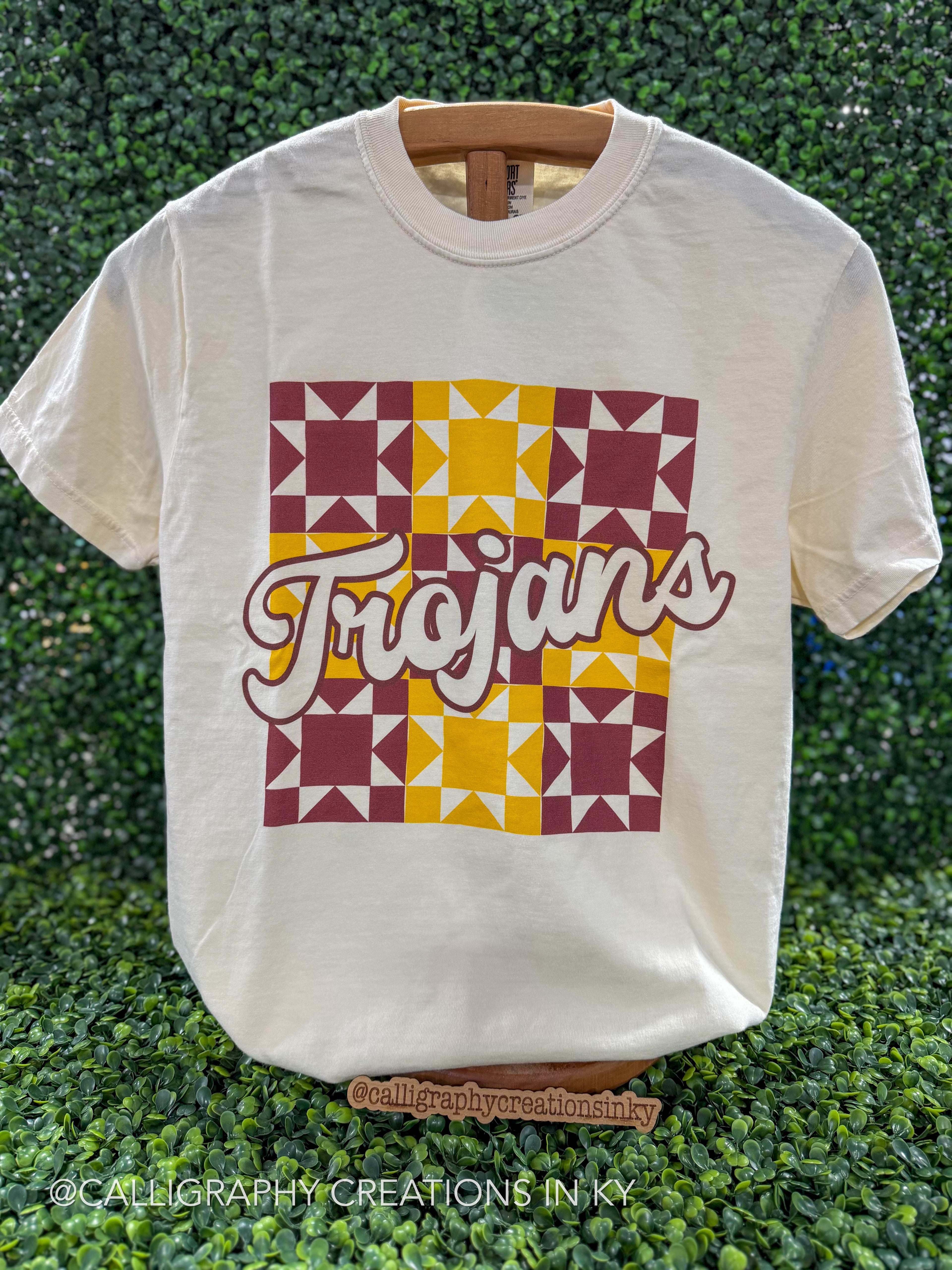 An off-white crewneck t-shirt with a retro-style geometric block print in maroon and yellow, featuring the word 'Trojans' in white script across the center. The shirt is displayed on a wooden hanger against a green hedge background.