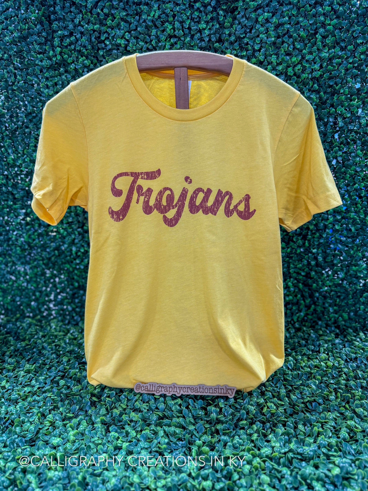 A yellow short-sleeve t-shirt with 'Trojan' written across the chest in a glittery dark red script, displayed on a hanger against a green textured background.
