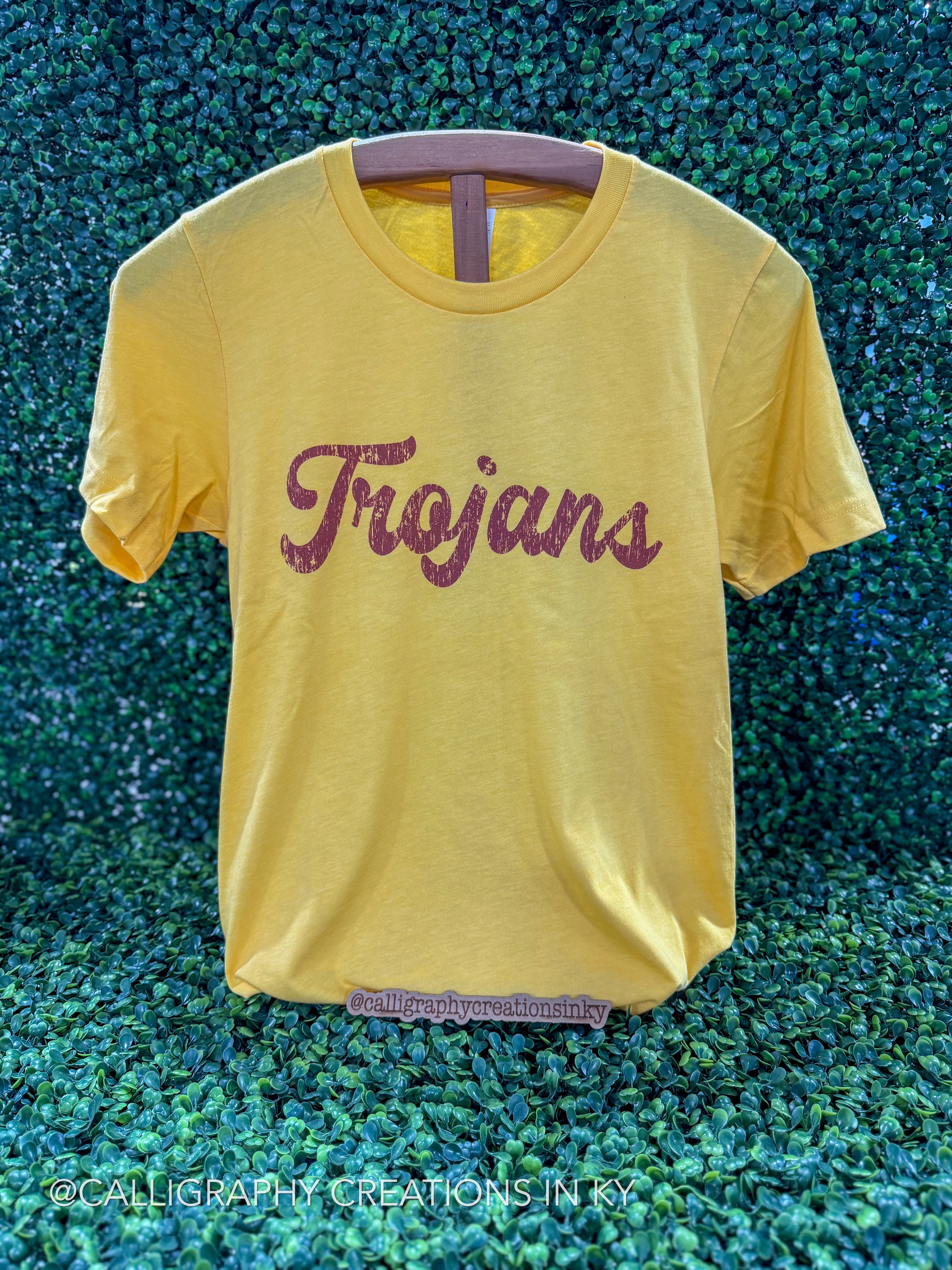 A yellow short-sleeve t-shirt with 'Trojan' written across the chest in a glittery dark red script, displayed on a hanger against a green textured background.