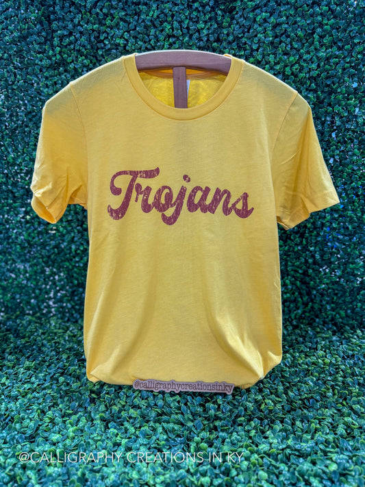 A yellow short-sleeve t-shirt with 'Trojan' written across the chest in a glittery dark red script, displayed on a hanger against a green textured background.