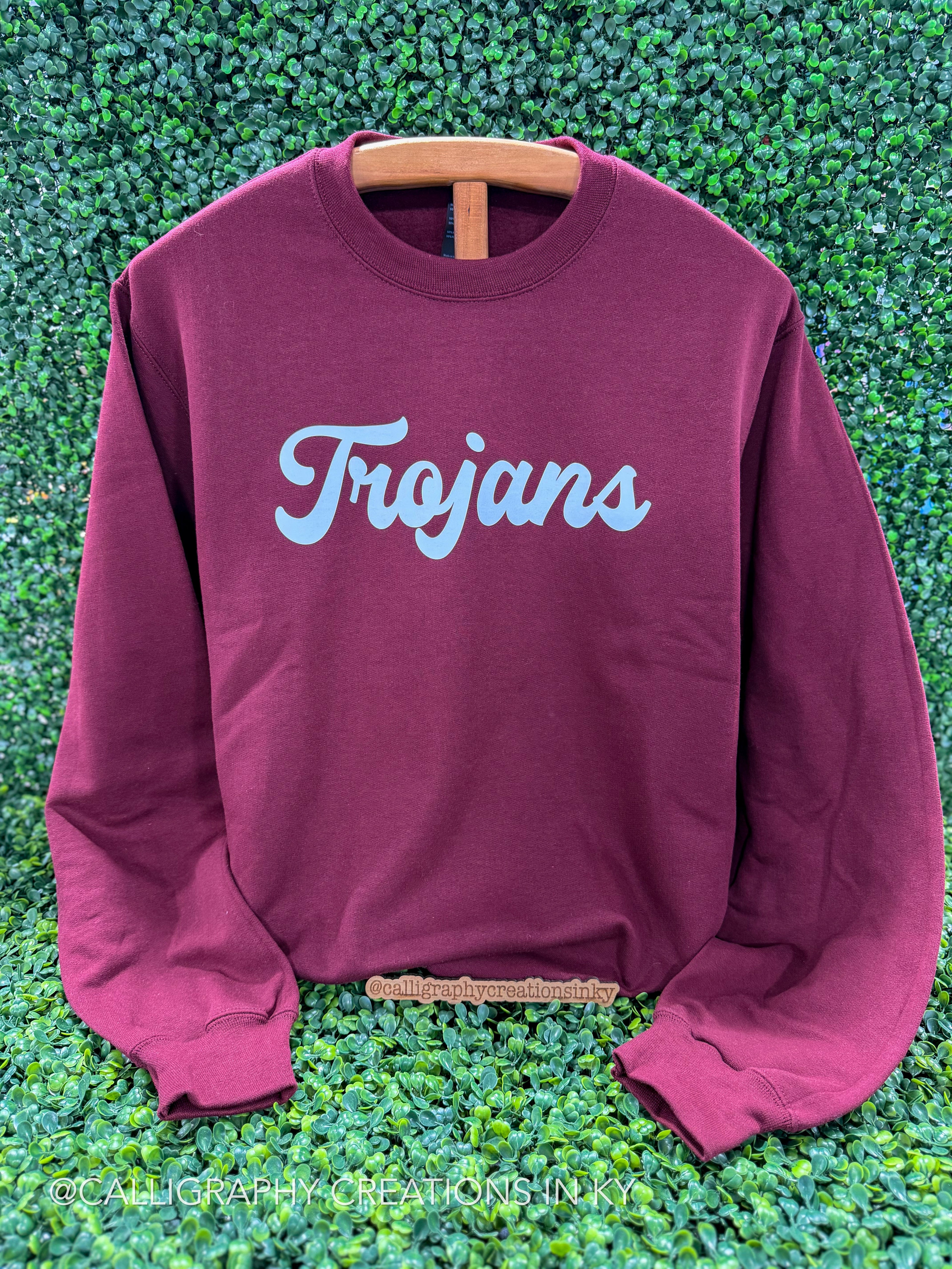 A maroon crewneck sweatshirt with 'Trojans' printed in light blue script across the chest, displayed on a wooden hanger against a green leafy background.